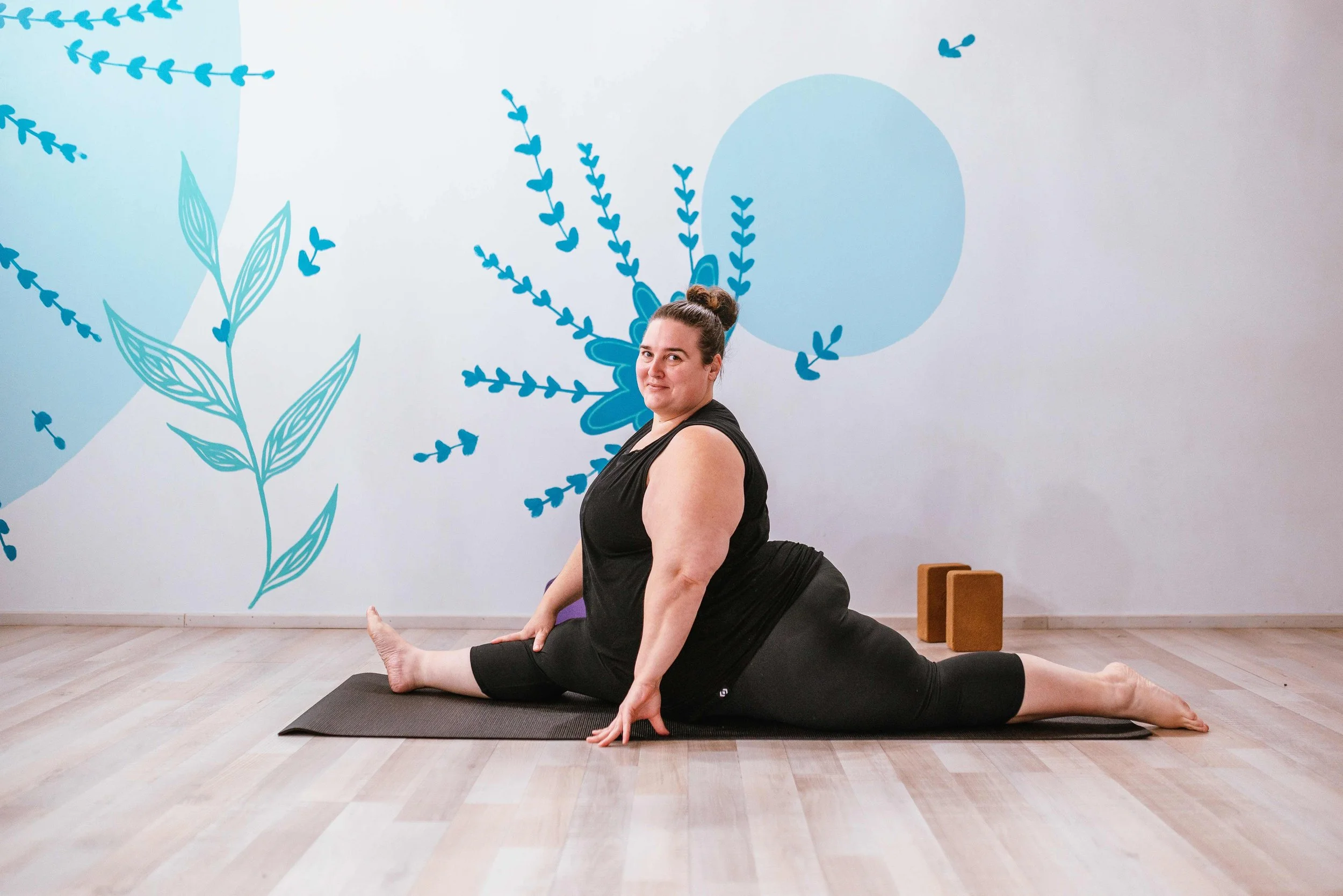A woman lying on her back on a yoga mat, with knees drawn towards her chest and hands holding her shins, in a yoga studio with wooden floors and a large blue ornamental wall mural. The studio has ambient lighting, a floor-standing lamp, and decorative objects like salt lamps and plants.