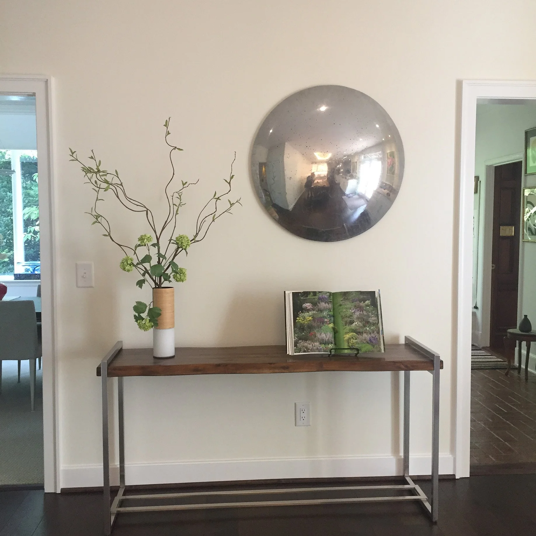 A modern hallway with a wooden and metal console table, a green floral book open on the table, a tall vase with branches and green flowers, a large circular reflective mirror on the wall, and two doorways on either side leading to other rooms.