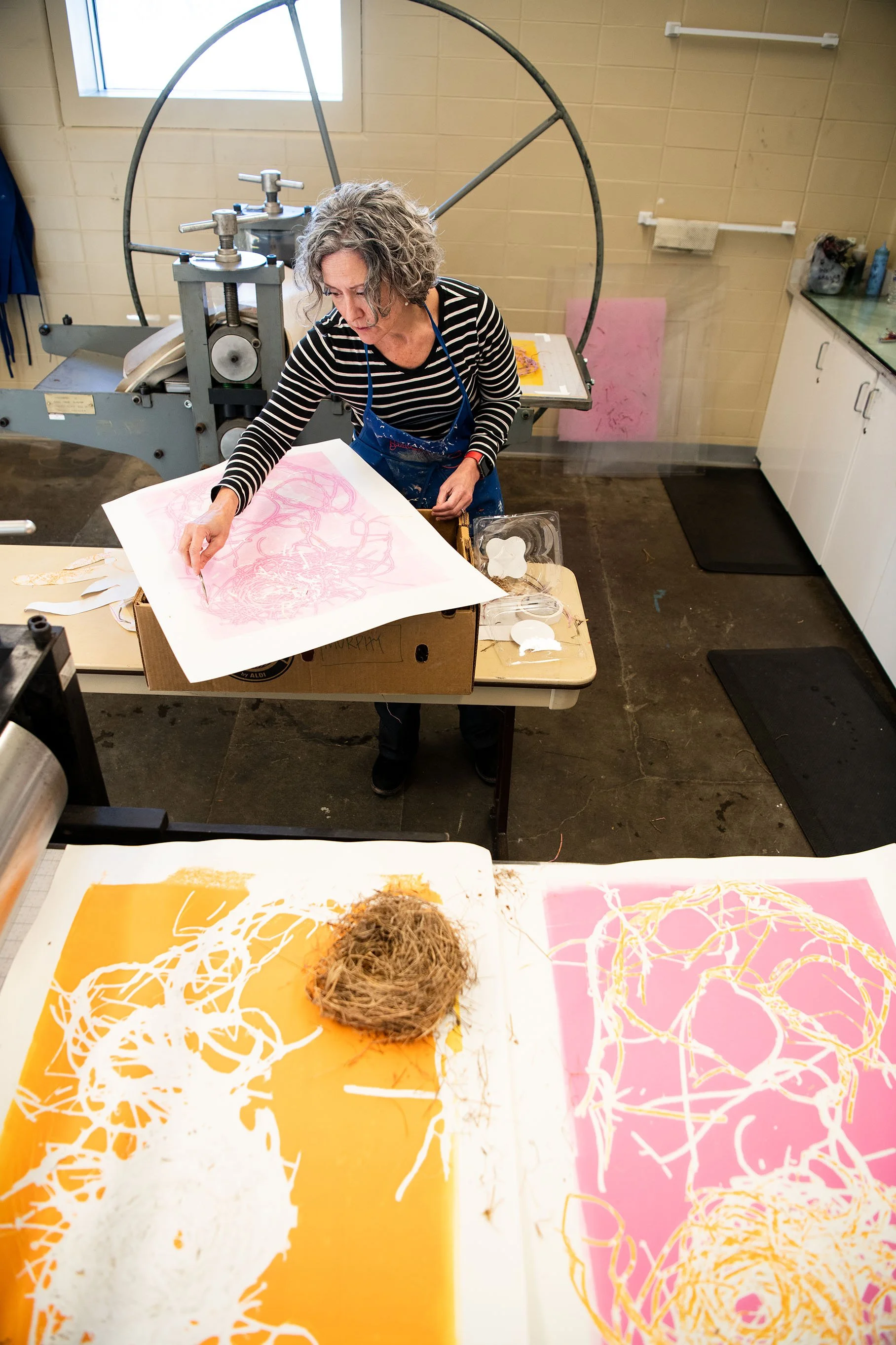 An artist working on a large printmaking project in a studio, with colorful artworks and art supplies on the table.