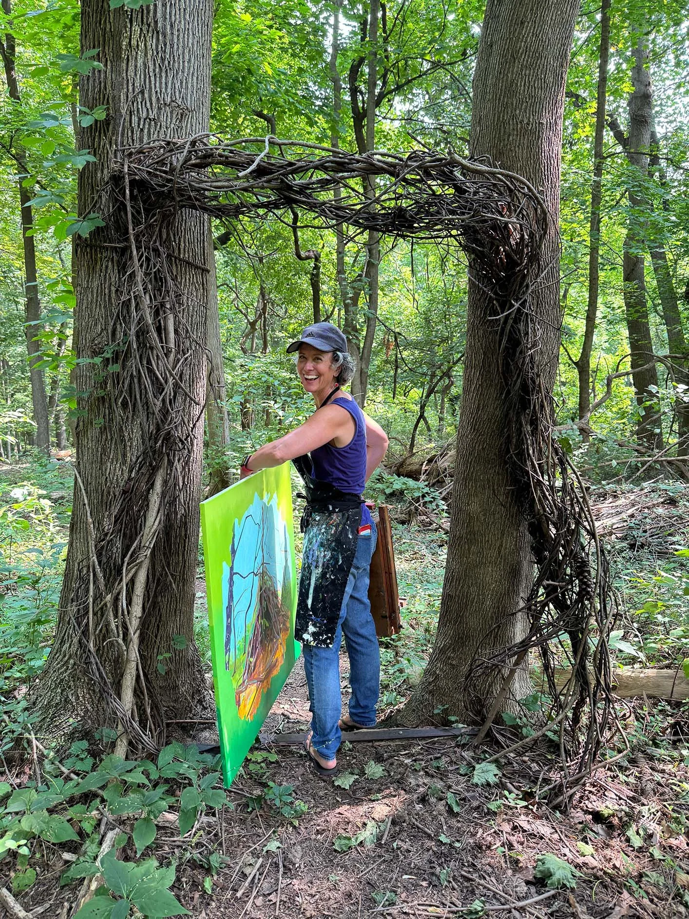 Artist, Deirdre Murphy, smiling in a forest, holding a colorful painting, standing under an arch made of intertwined tree branches and vines.