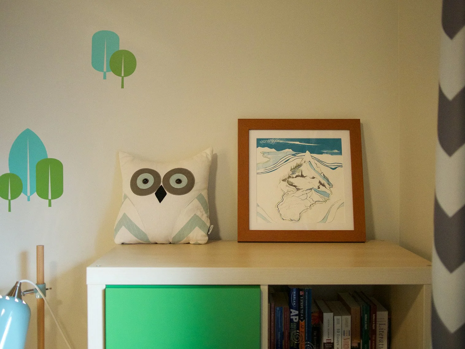 Decorative owl pillow and framed mountain landscape artwork on a shelf in a room with tree wall decals.