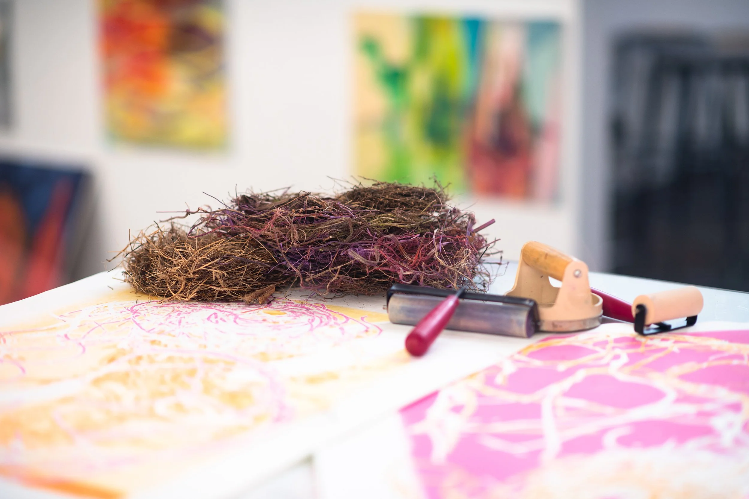 Art supplies on a table, including a bundle of dried plant stems, a stapler, and a pink-handled roller, with colorful drawings or paintings in the background.