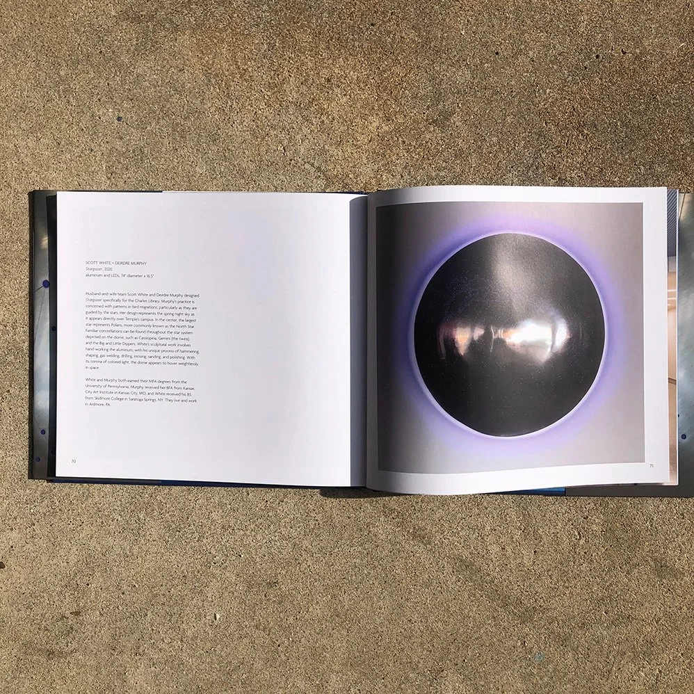 Open book on a sandy surface showing a photograph of an art piece featuring a black circular object with a purple glow and reflective surface.
