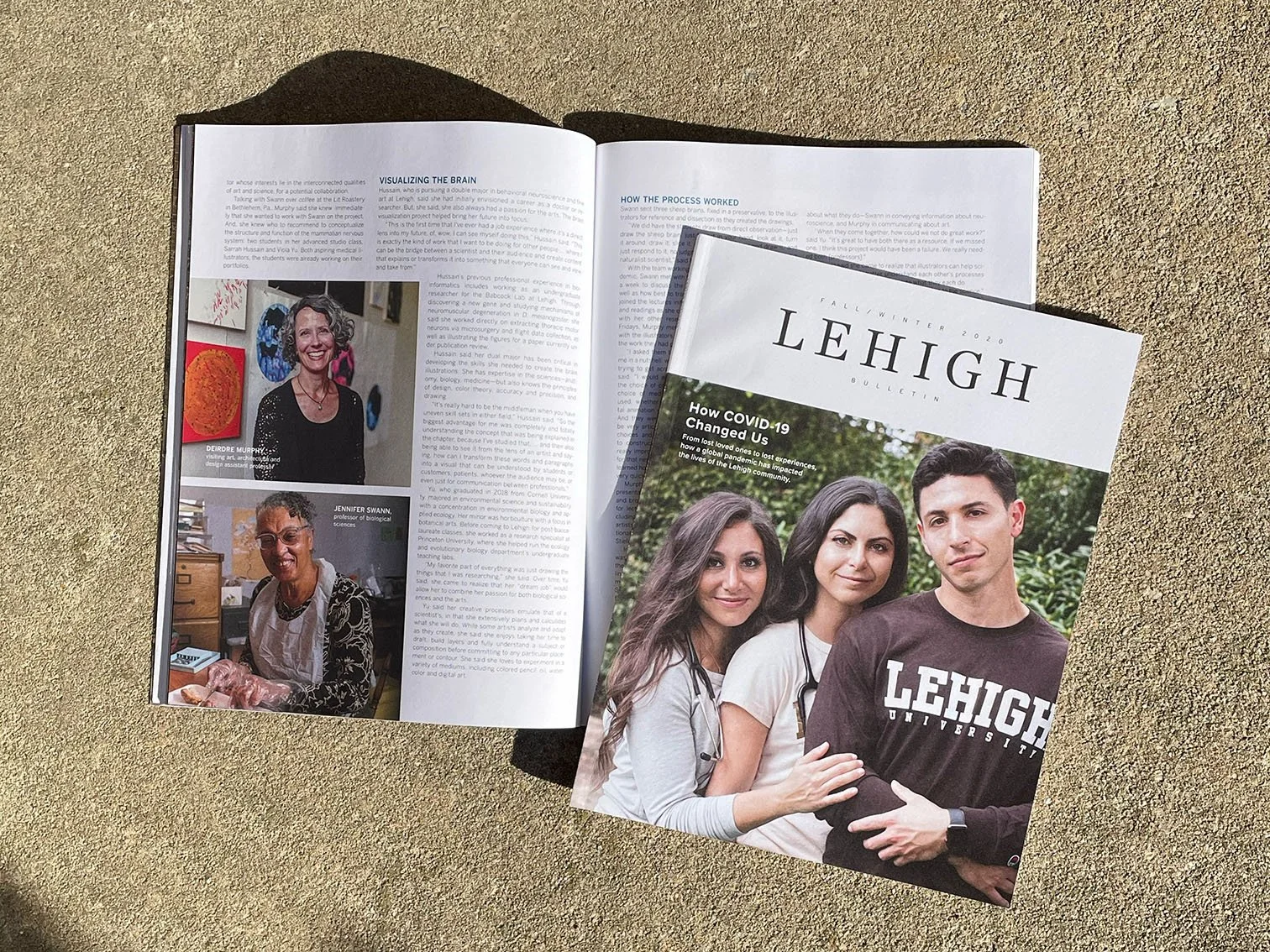 Open magazine on sand with cover showing three young adults, two women and one man, outdoors, some reading material inside.