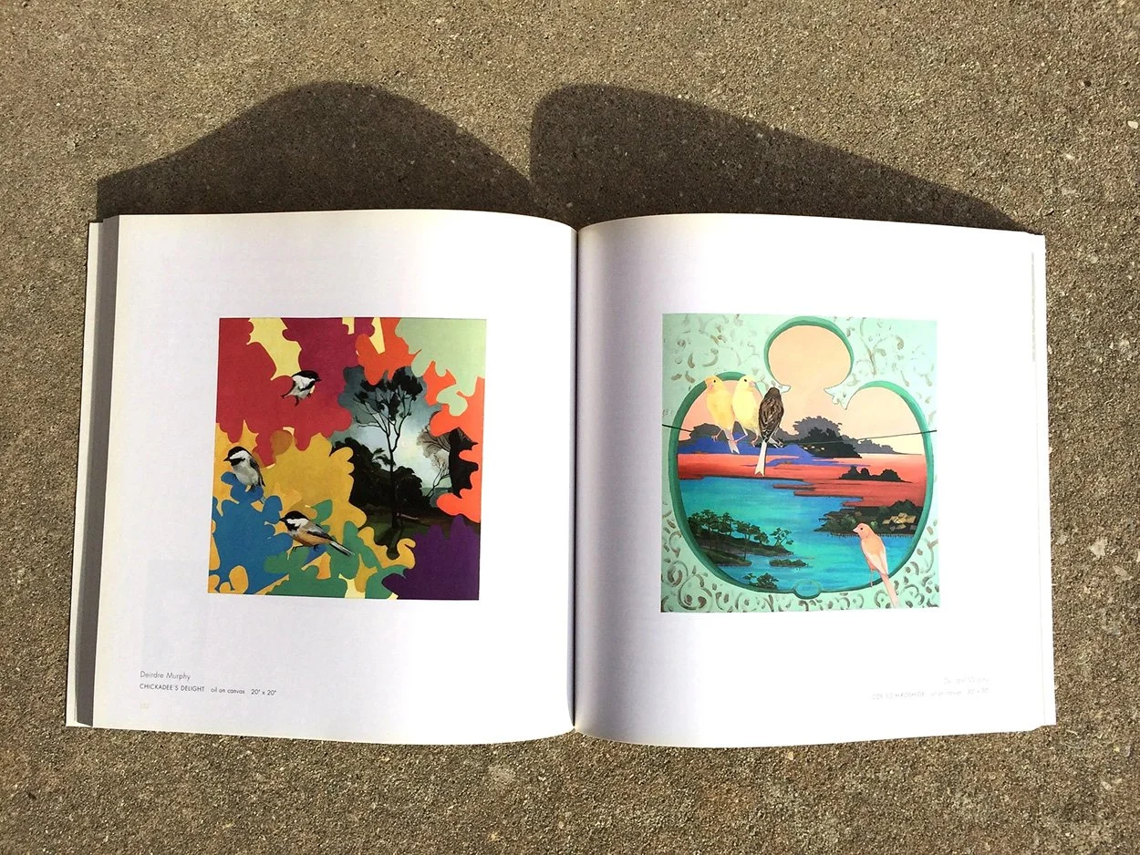 Open art book displaying two colorful landscape paintings of birds and nature scenes, with the book resting on a rough, sand-colored surface.