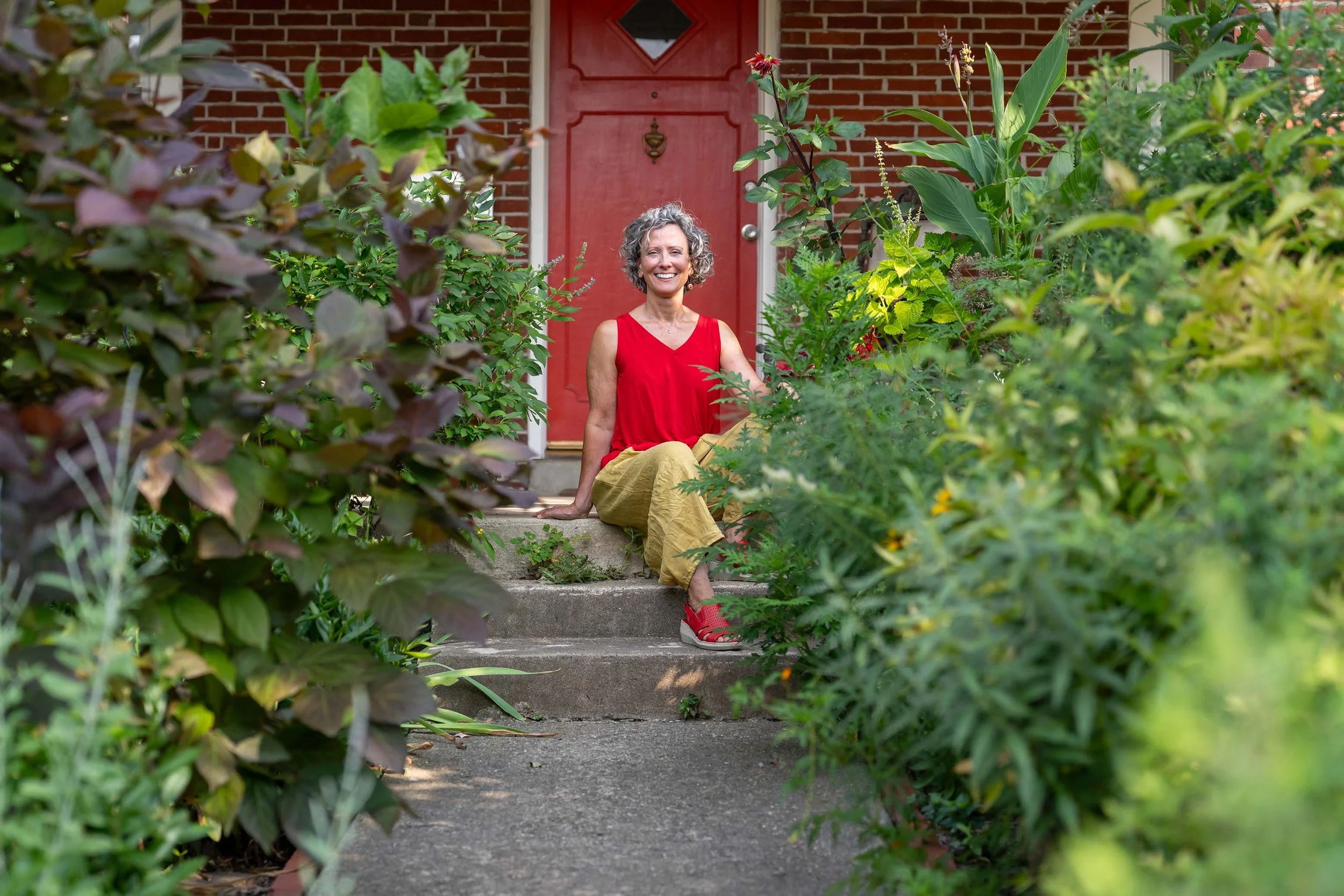 Artist Deirdre Murphy - a woman with curly gray hair smiling, sitting on steps in front of a red door, surrounded by lush green plants and bushes.