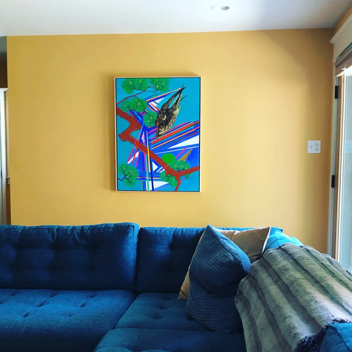Living room with a yellow wall, blue couch with pillows, and a colorful framed artwork featuring a bird on a tree branch.