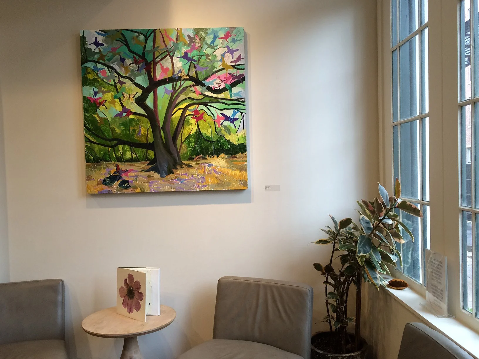 Colorful painting of a tree with pink, purple, green, and yellow leaves and flying birds hanging on a gallery wall next to a window with potted plant and seating area.