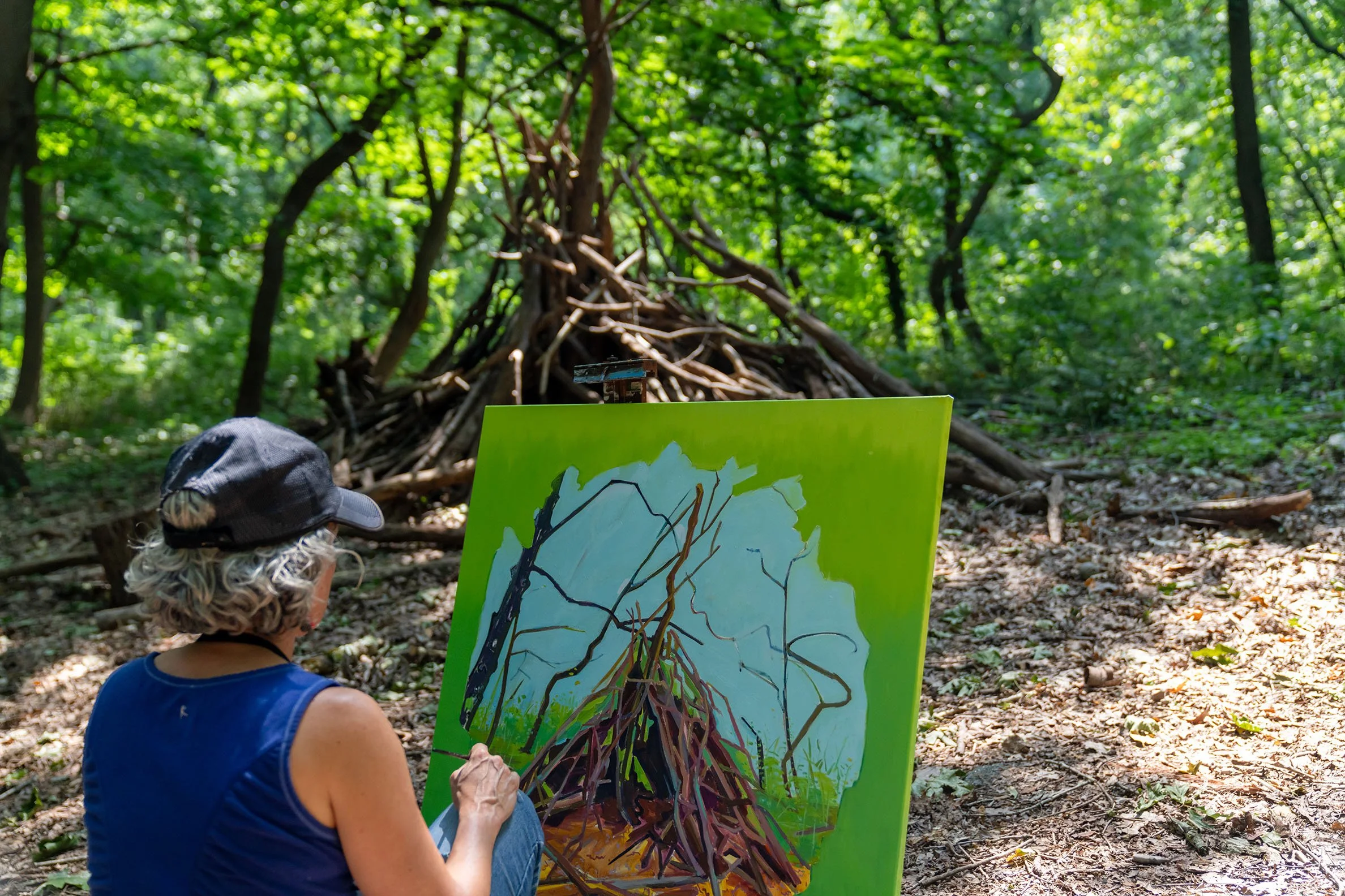 An artist creating a painting of a teepee-like structure made of sticks and branches in a forest.