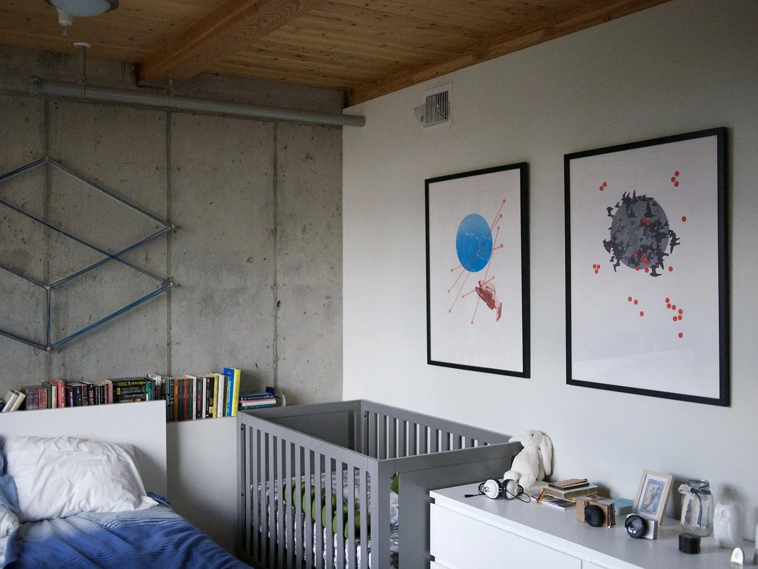A bedroom with a concrete wall and wooden ceiling, featuring two framed abstract art pieces on a white wall, a bed with white and blue bedding, a white dresser with various items including books, headphones, and framed pictures, and a gray crib besid