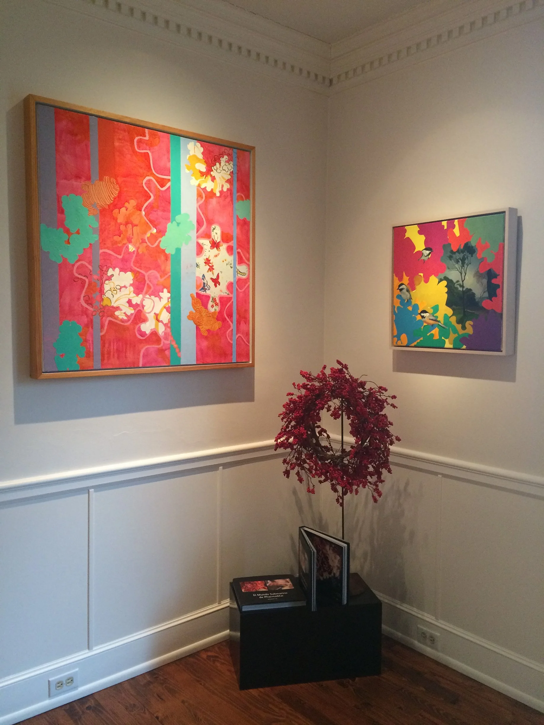 Art gallery display with two colorful abstract paintings on white walls and a small black pedestal holding books and a wreath made of pink flowers.