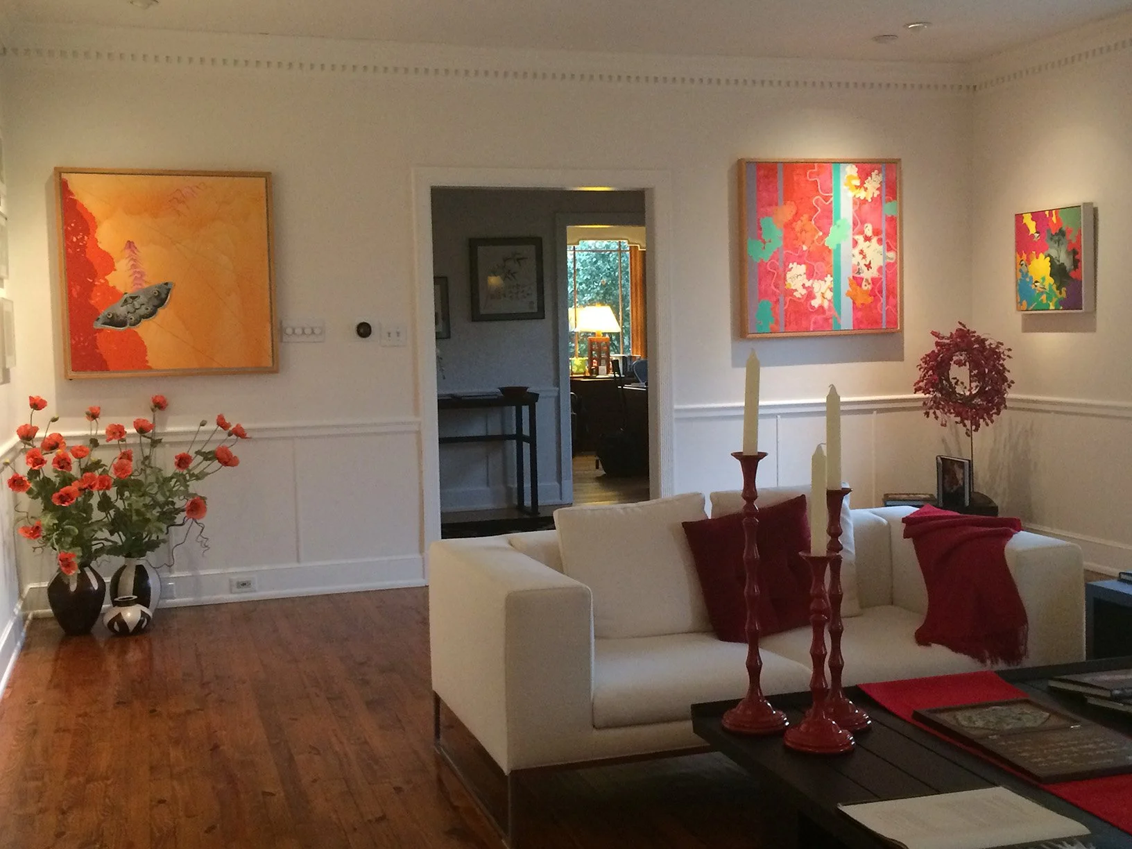 Living room with white walls, hardwood floor, decorated with colorful abstract paintings, large floral arrangements, and a white sofa with red pillows and throws. Candles on a black table in front of the sofa.
