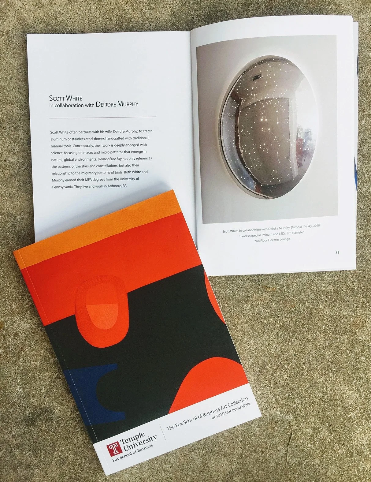 An open book displaying a page with text about Scott White in collaboration with Deirdre Murphy, and a photograph of a circular, hand-shaped aluminum and LED artwork titled "Dome of the Sky" on the right page. A folded booklet with orange, black, red