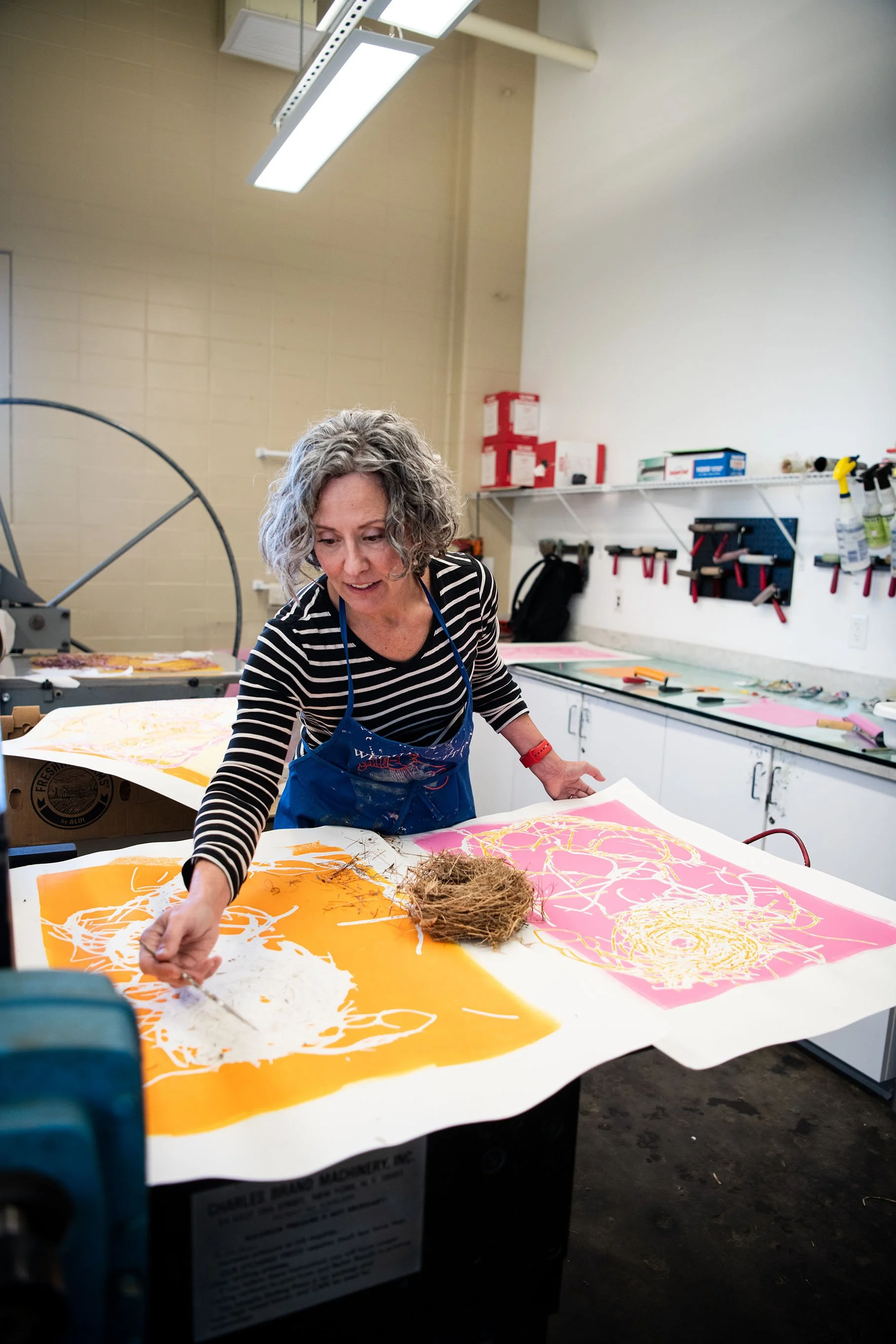 A woman with curly gray hair wearing a navy apron and striped shirt working on colorful prints in an art studio.