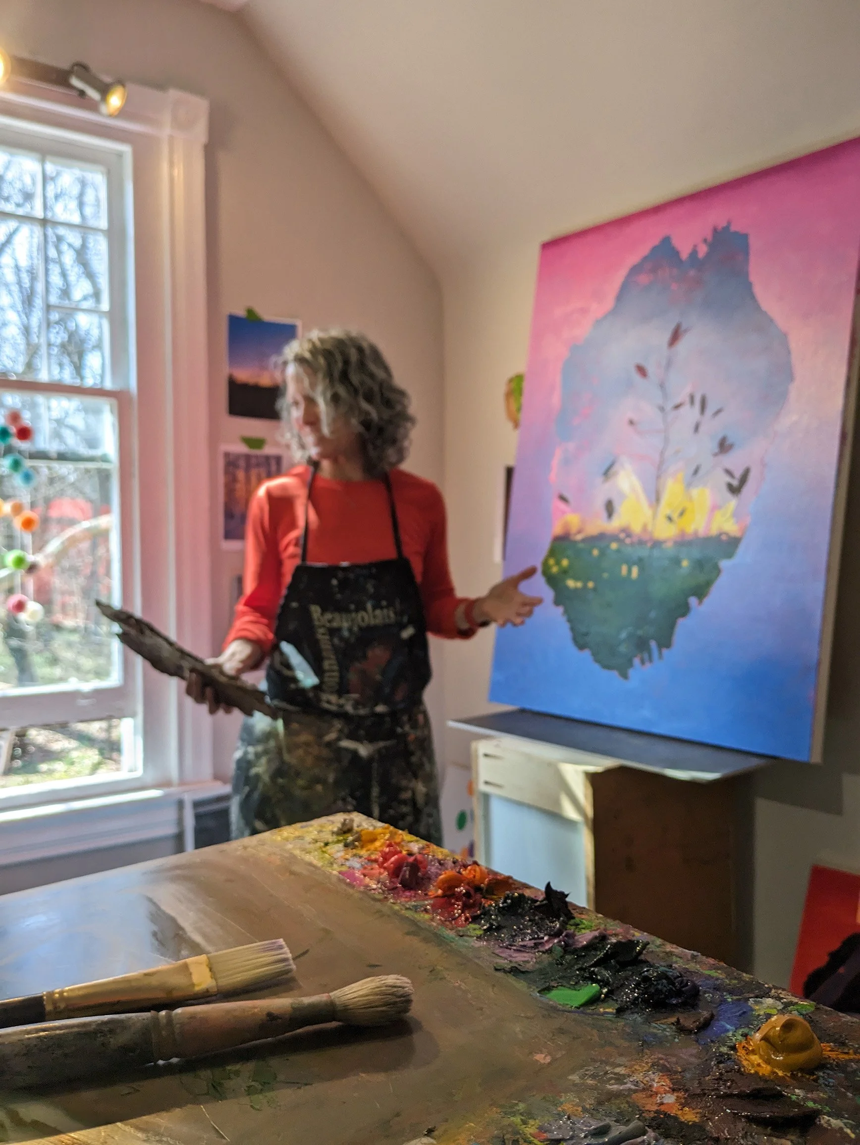An artist with curly gray hair wearing a red shirt and an apron, holding a palette, standing in front of a colorful painting of a mountain with a tree, inside an art studio with brushes and paint on a wooden table.