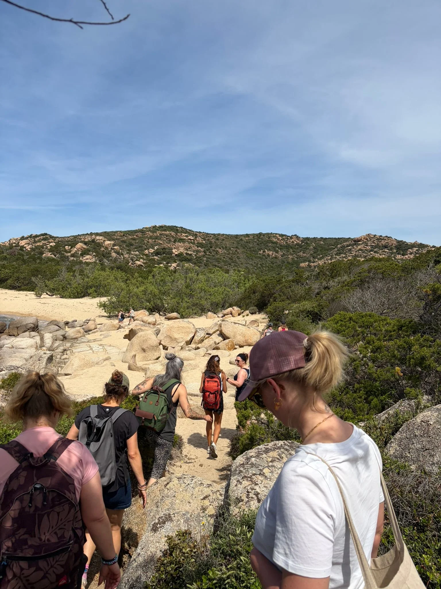 Groupe de personnes marchant sur un sentier de randonnée dans une région rocheuse avec des collines et une végétation dense, ciel bleu en arrière-plan.