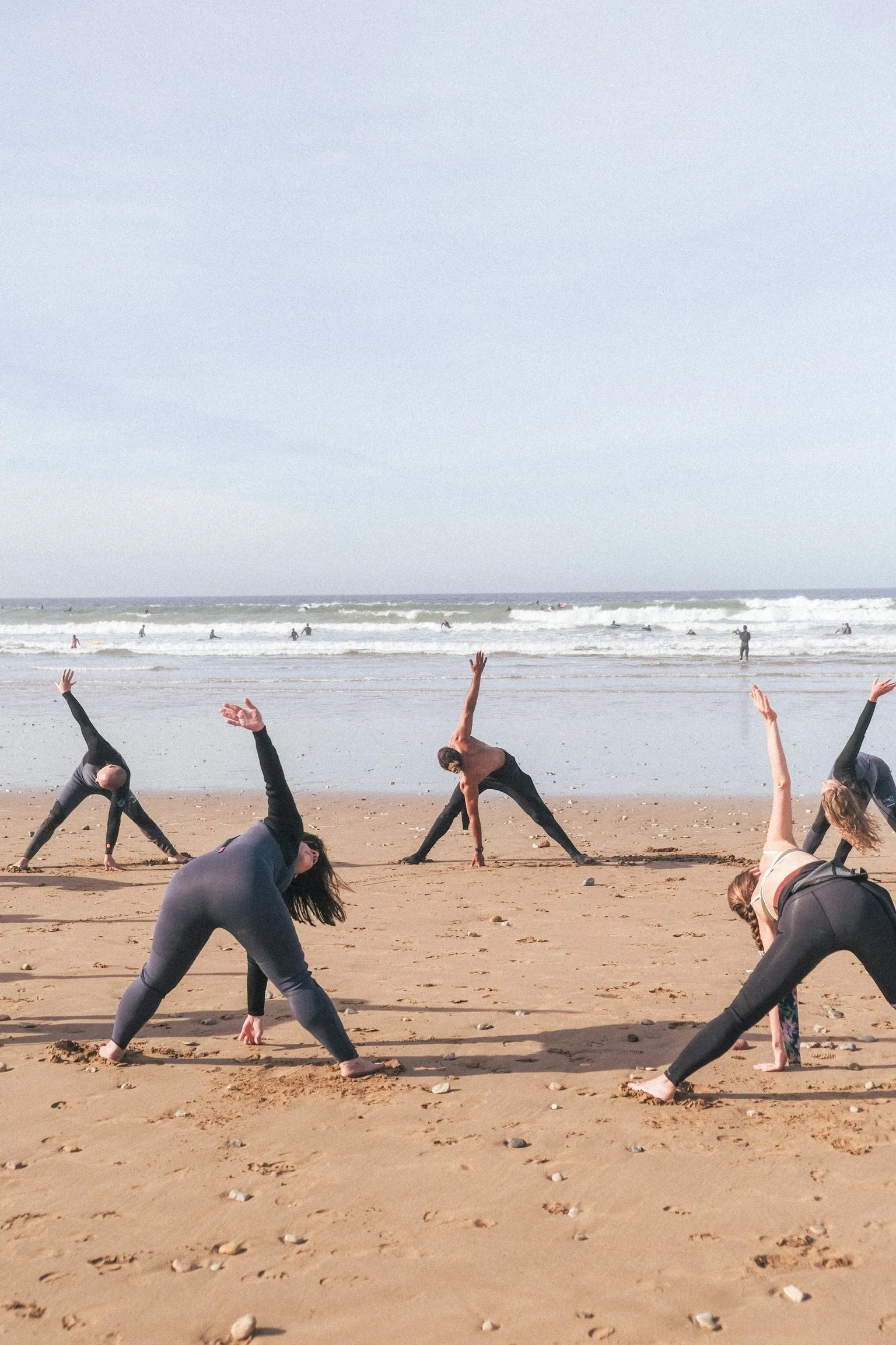 Échauffement collectif sur la plage face à l’océan, avec un groupe en combinaison préparant le corps avant une session de surf au Maroc.
