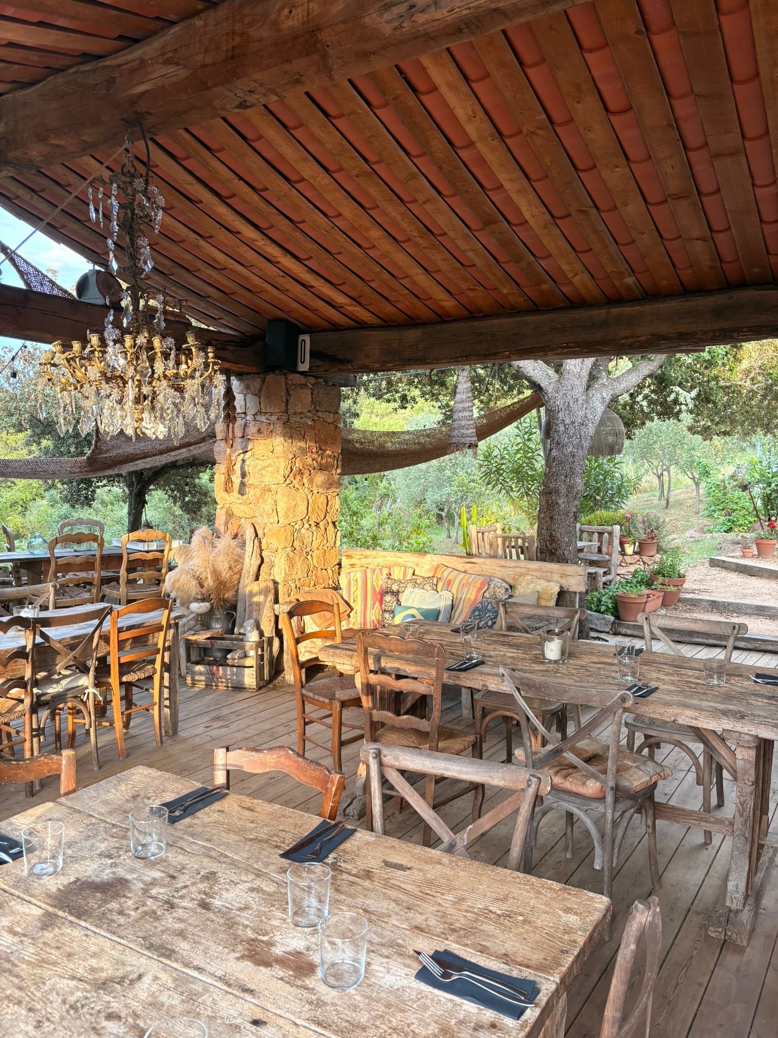 Une terrasse en bois avec des tables en bois rustiques, des verres, des couverts et des bougies, entourée d'arbres et de plantes, avec un plafond en bois et une décoration vintage, dans un cadre naturel calme et verdoyant.