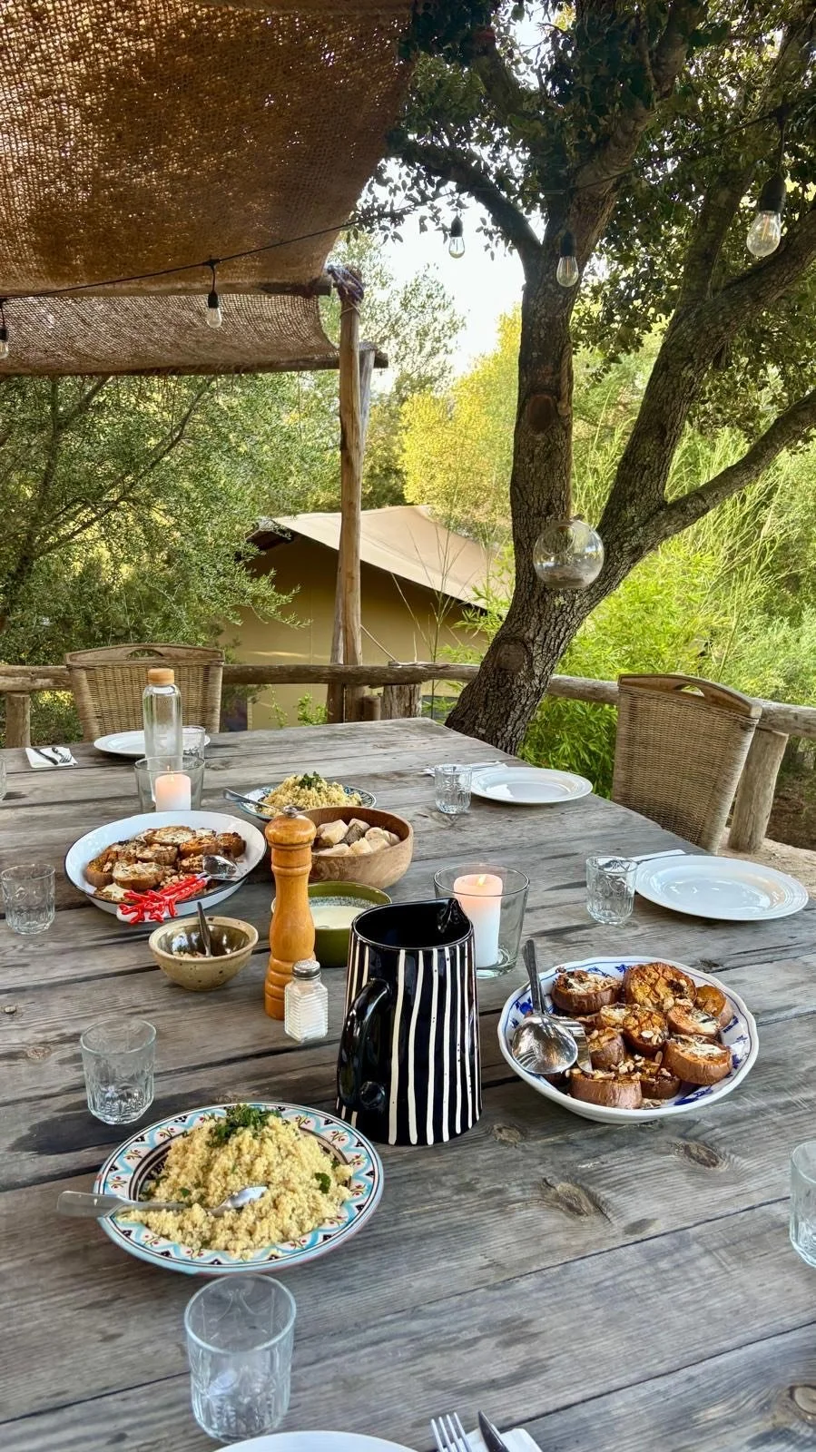 Repas de style campagnard avec variétés de plats, assiettes, verres et bougies, en plein air entouré d'arbres.