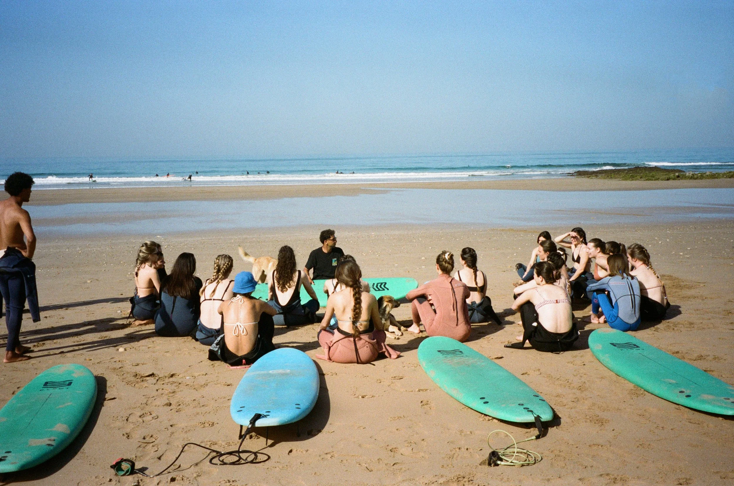 Groupe de personnes assises sur la plage avec des planches de surf, vue sur la mer et des surfeurs dans l'eau.