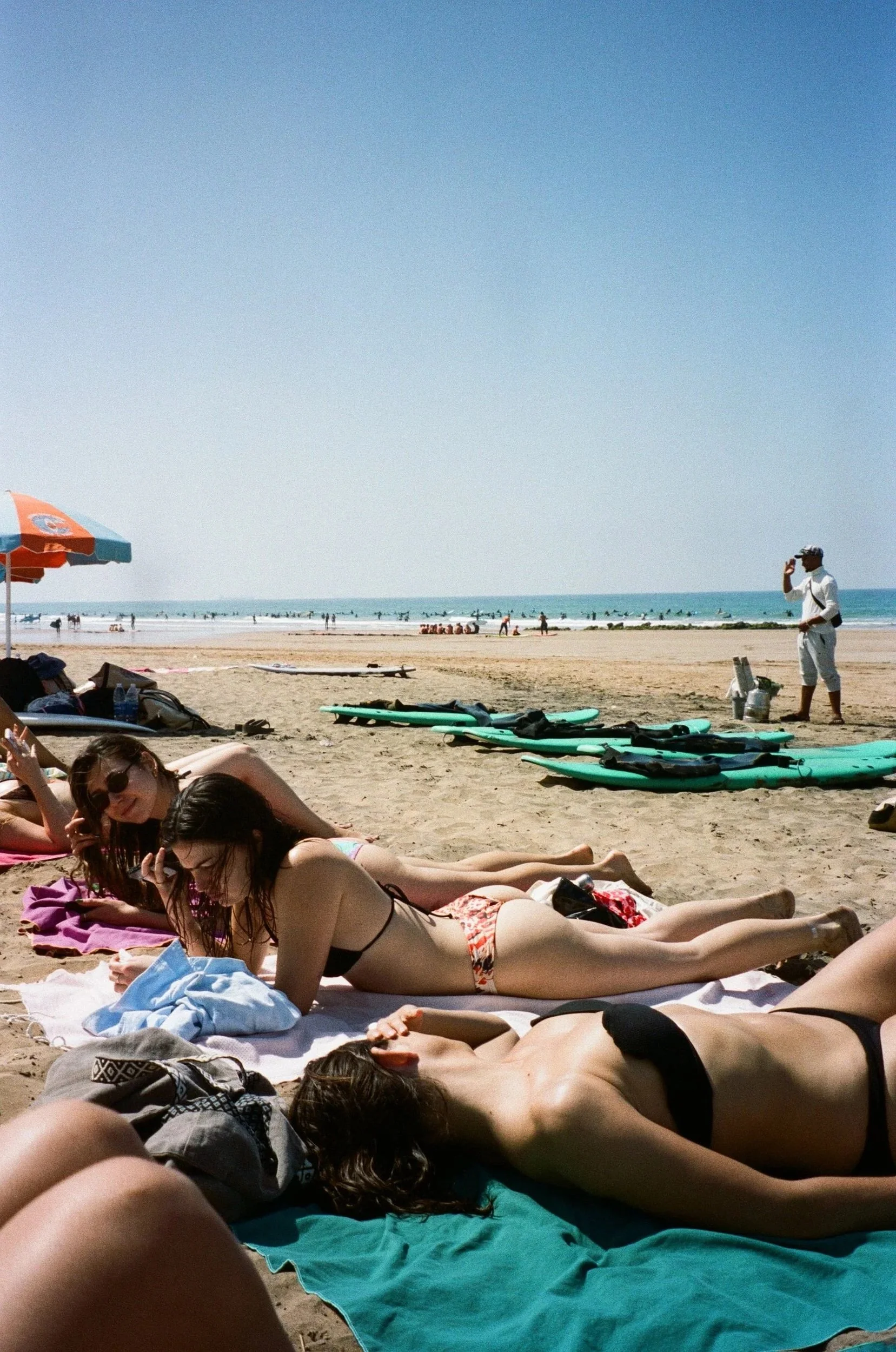 Des personnes allongées sur la plage, certains en maillot de bain, profitant du soleil. En arrière-plan, on voit la mer avec des surfers et un homme en tenue décontractée parlant au téléphone, sous un parasol orange et blanc.