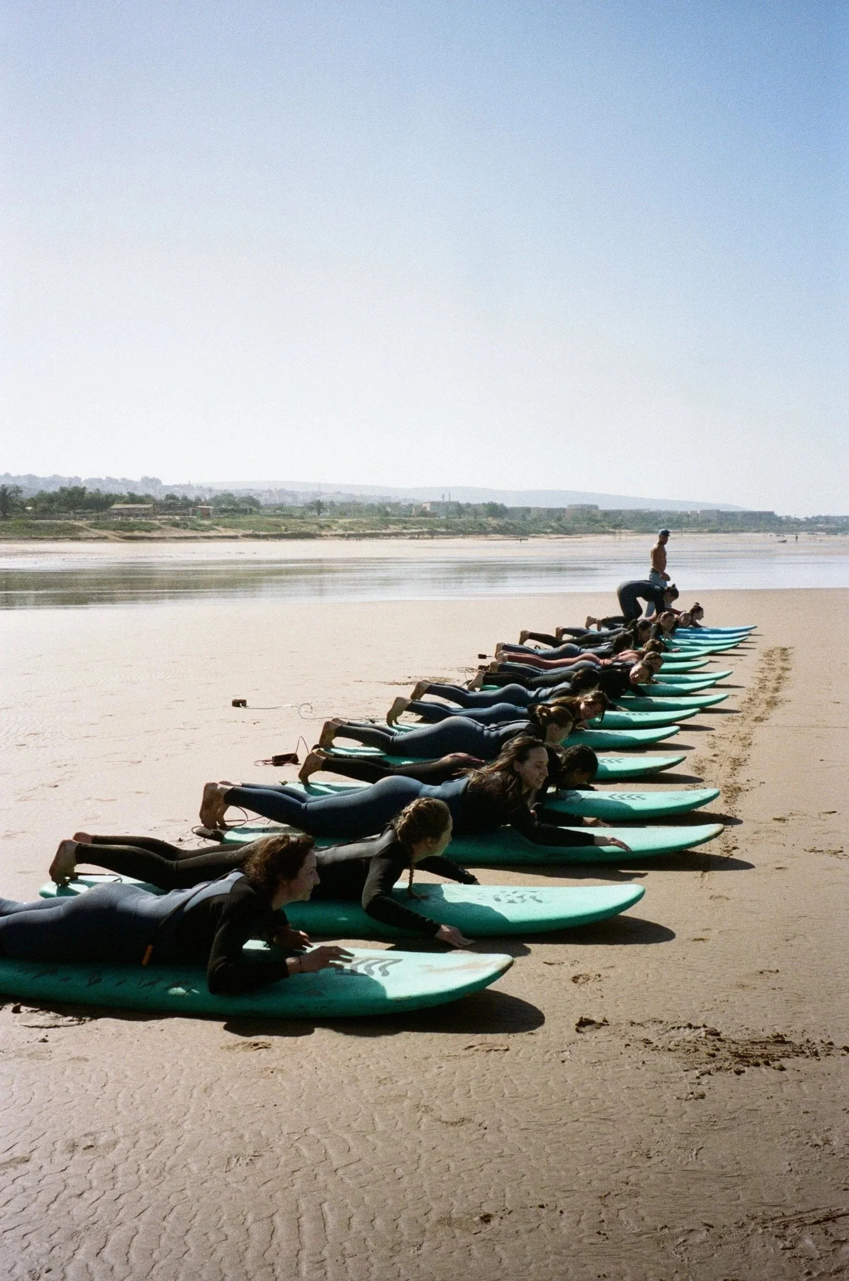 Groupe de personnes en position de planche sur une plage, avec un instructeur debout sur un seul d'eux, pratiquant probablement le yoga ou le Pilates, avec la mer en arrière-plan.