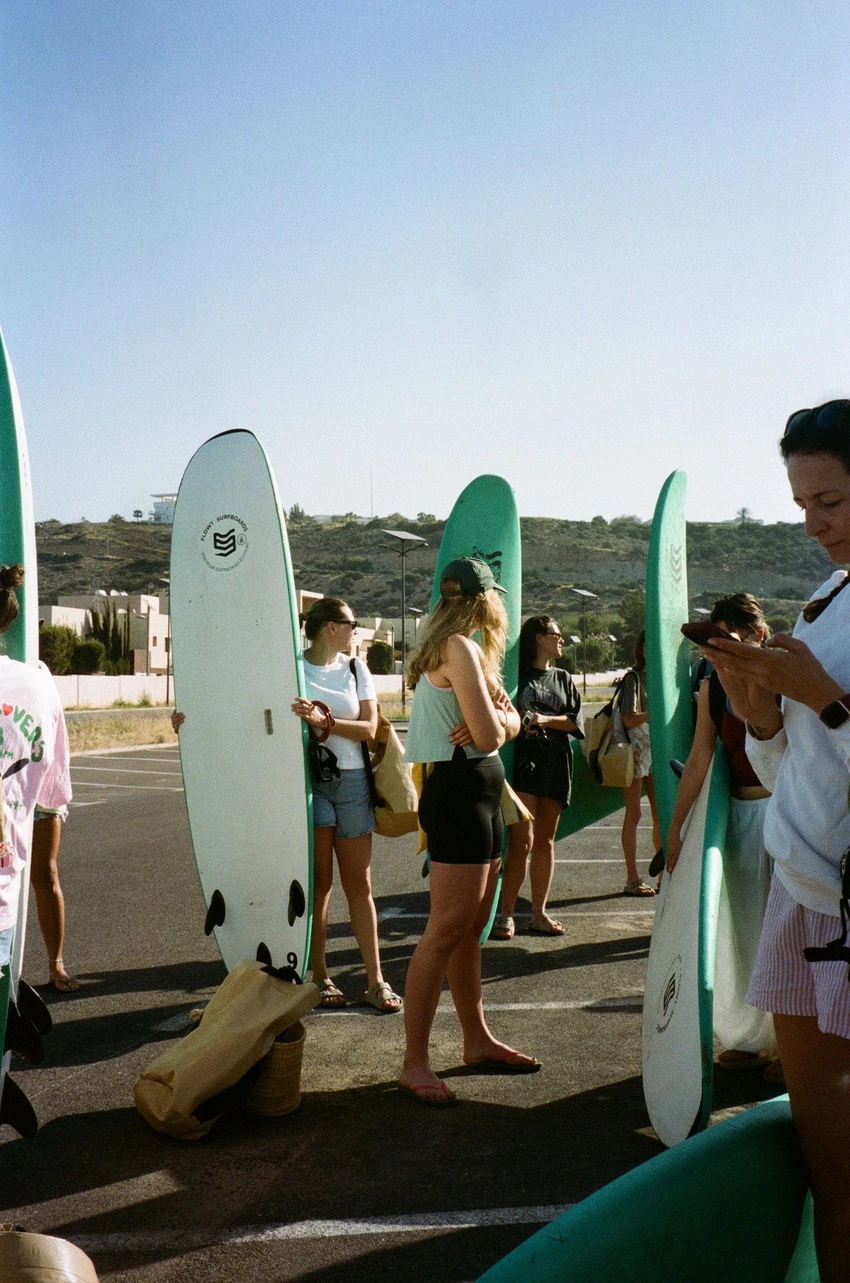 Groupe de personnes debout avec des planches de surf vertes dans un parking extérieur, certains utilisant leur téléphone, sous un ciel clair.