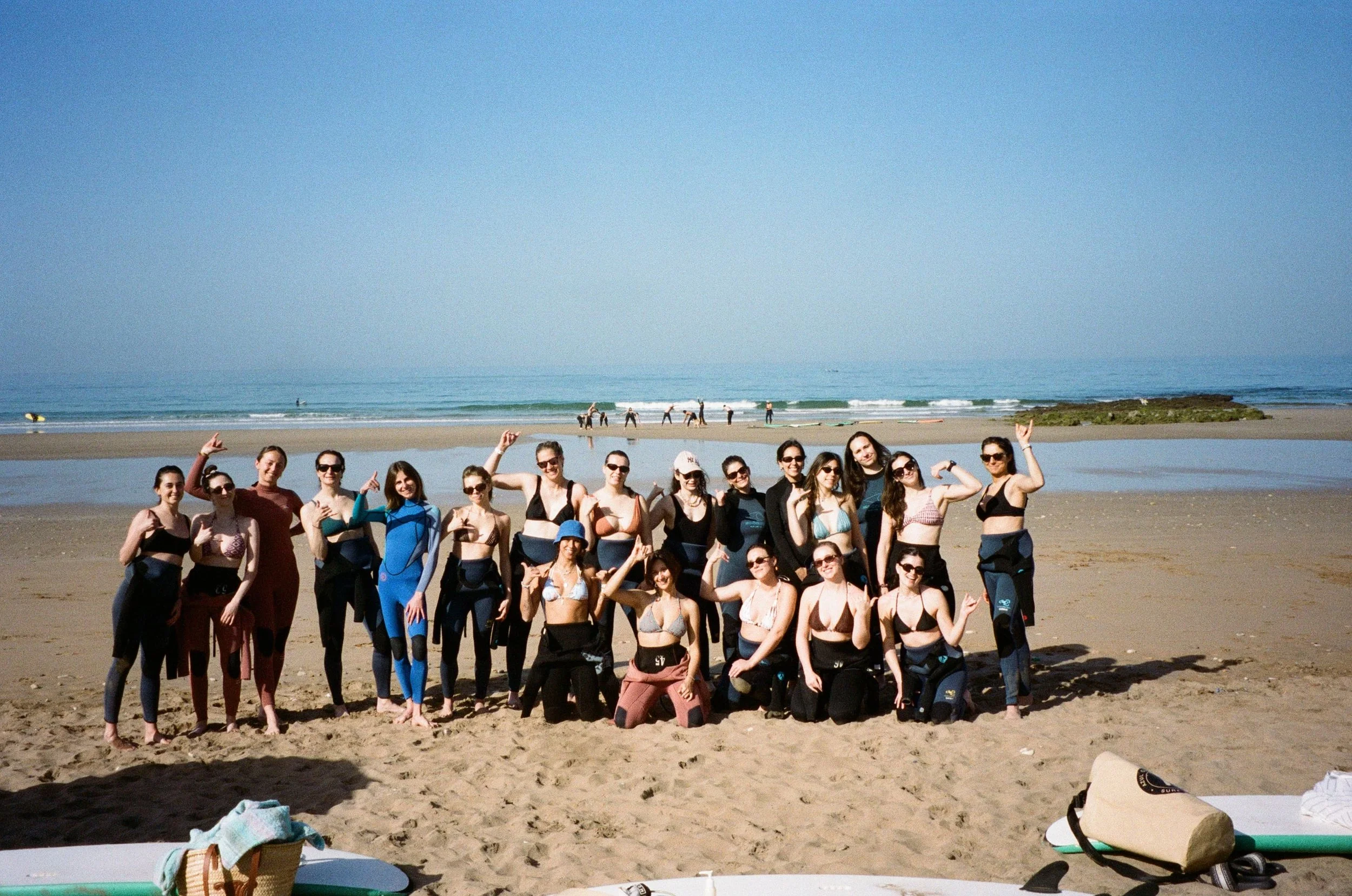 Groupe de personnes posant sur la plage avec la mer en arrière-plan, certaines portent des vêtements de sport ou maillots de bain, probablement après une séance de surf ou un autre sport aquatique.