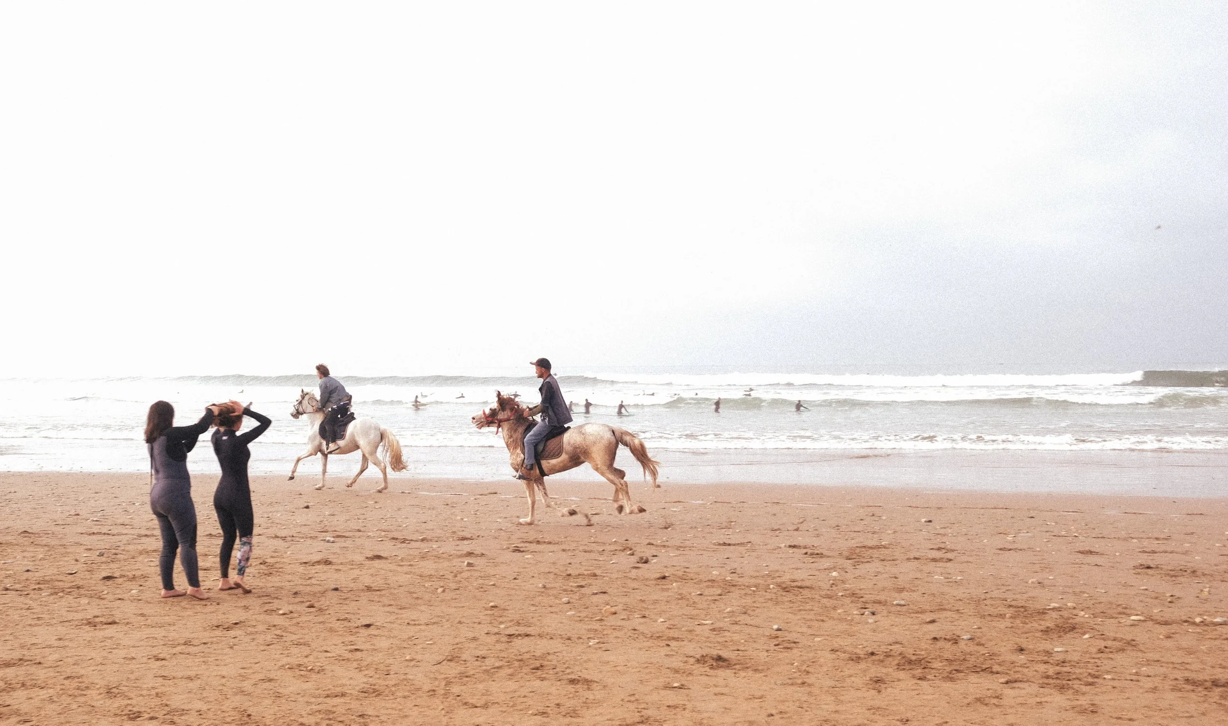 Balade à cheval sur une plage marocaine, avec l’océan et les surfeurs en arrière-plan, dans une ambiance douce et dépaysante.