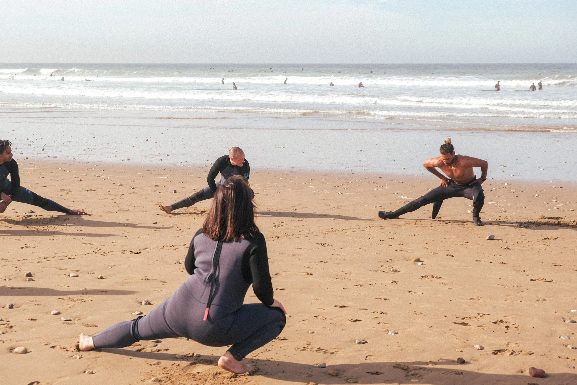 Échauffement collectif sur la plage avant une session de surf, face aux vagues et à l’océan, dans une ambiance sportive et conviviale.