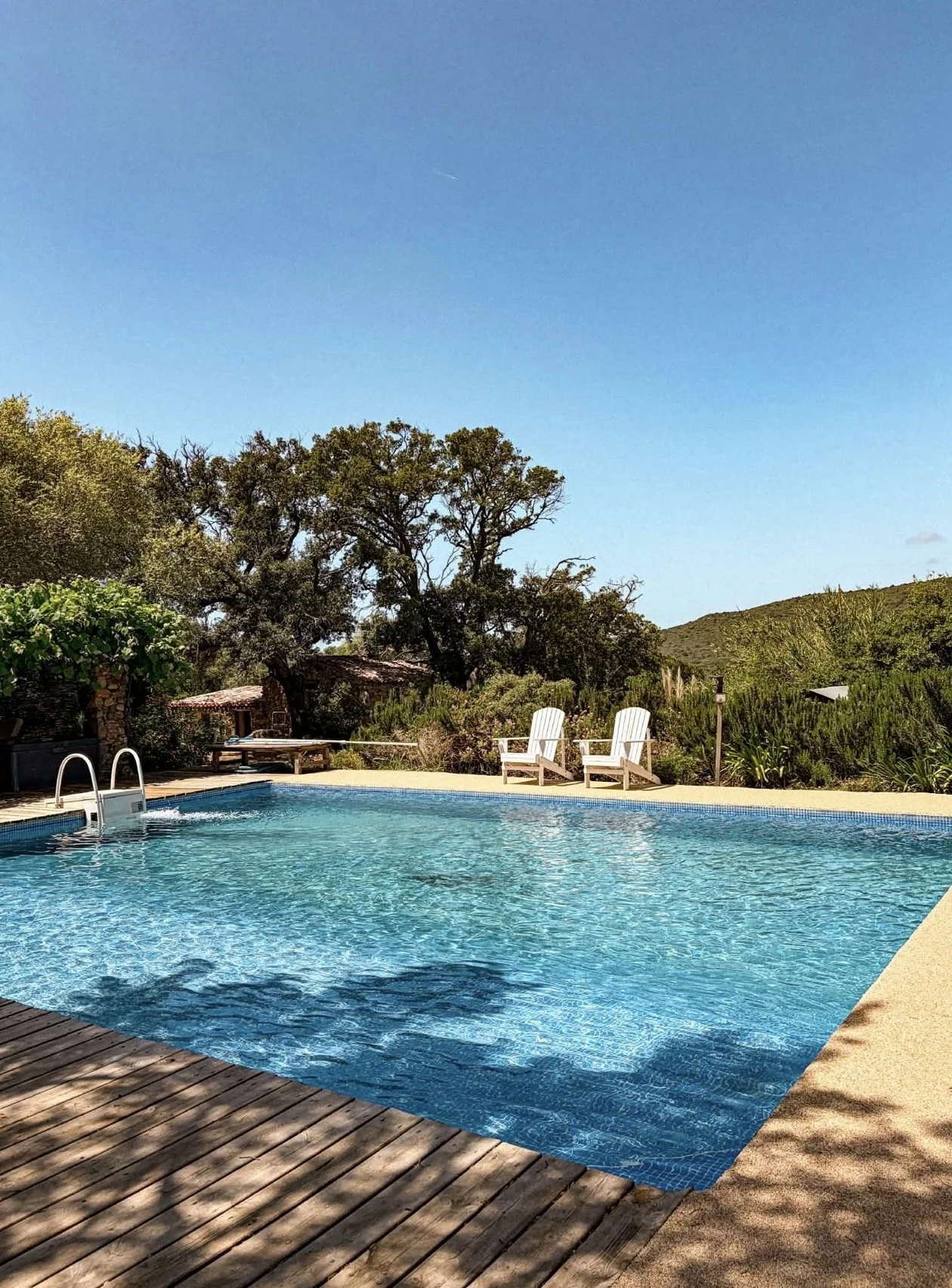 Une piscine extérieure avec deux chaises longues blanches, entourée de végétation et de collines, sous un ciel bleu clair.