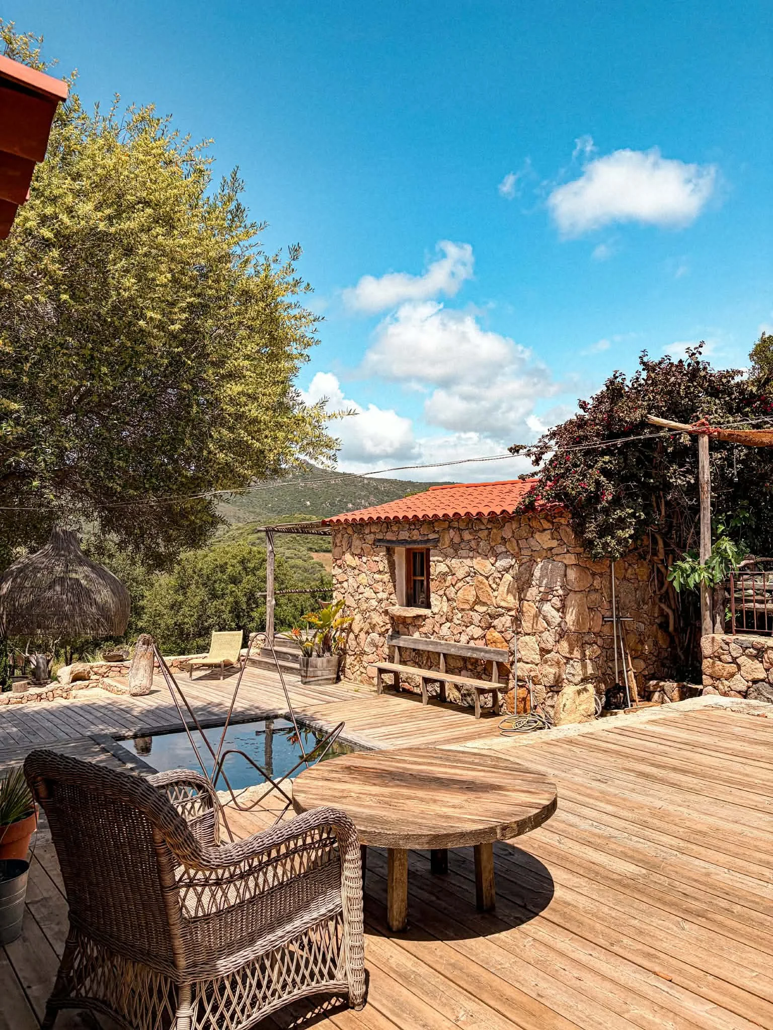 Terrasse en bois avec chaises et table en rotin, petite piscine, maison en pierre avec tuiles oranges, arbres et ciel bleu avec quelques nuages.