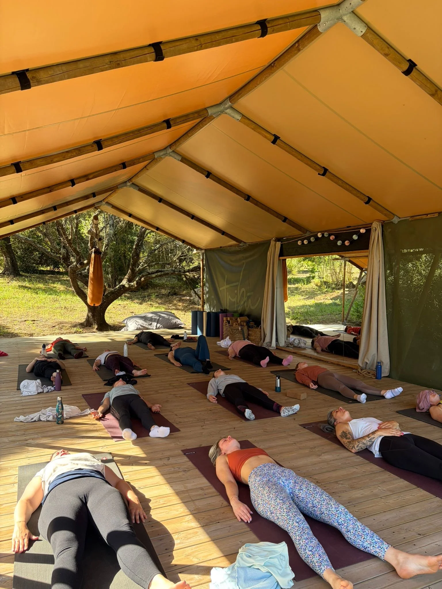 Groupe de personnes faisant du yoga en position allongée sur des tapis dans une tente en plein air, sous un toit en tissu orange, avec des arbres en arrière-plan.