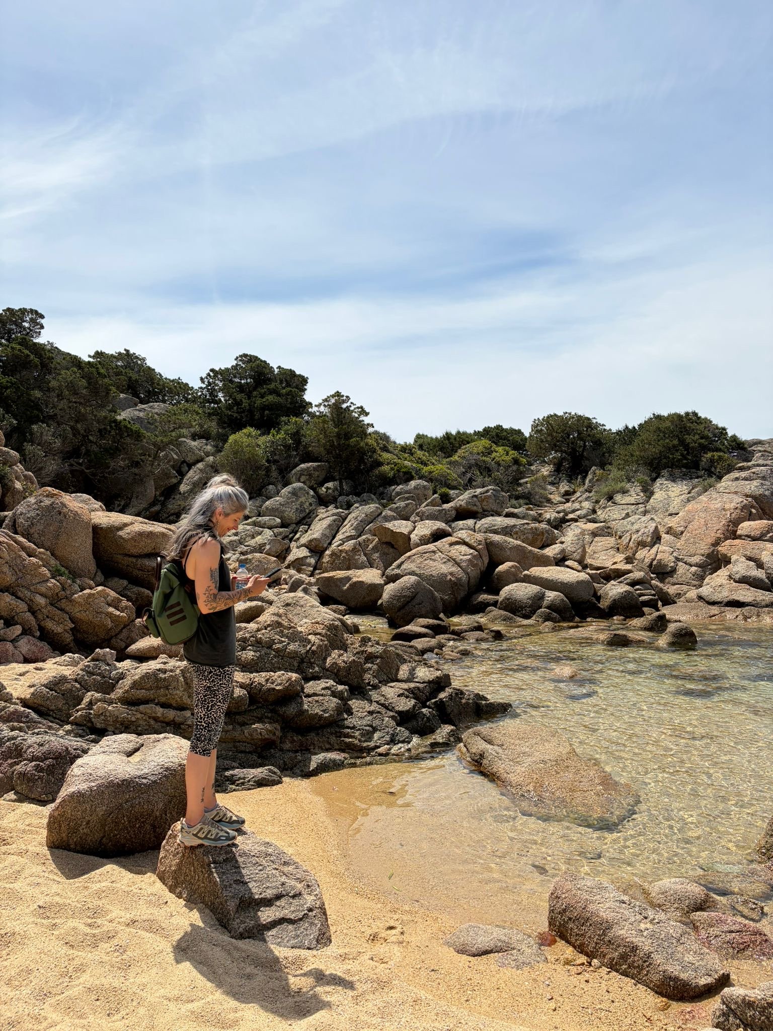 Femme à cheveux gris, portant un sac à dos vert, des vêtements décontractés, se tenant sur des rochers au bord de l'eau dans un paysage naturel avec des arbres et un ciel bleu avec quelques nuages.
