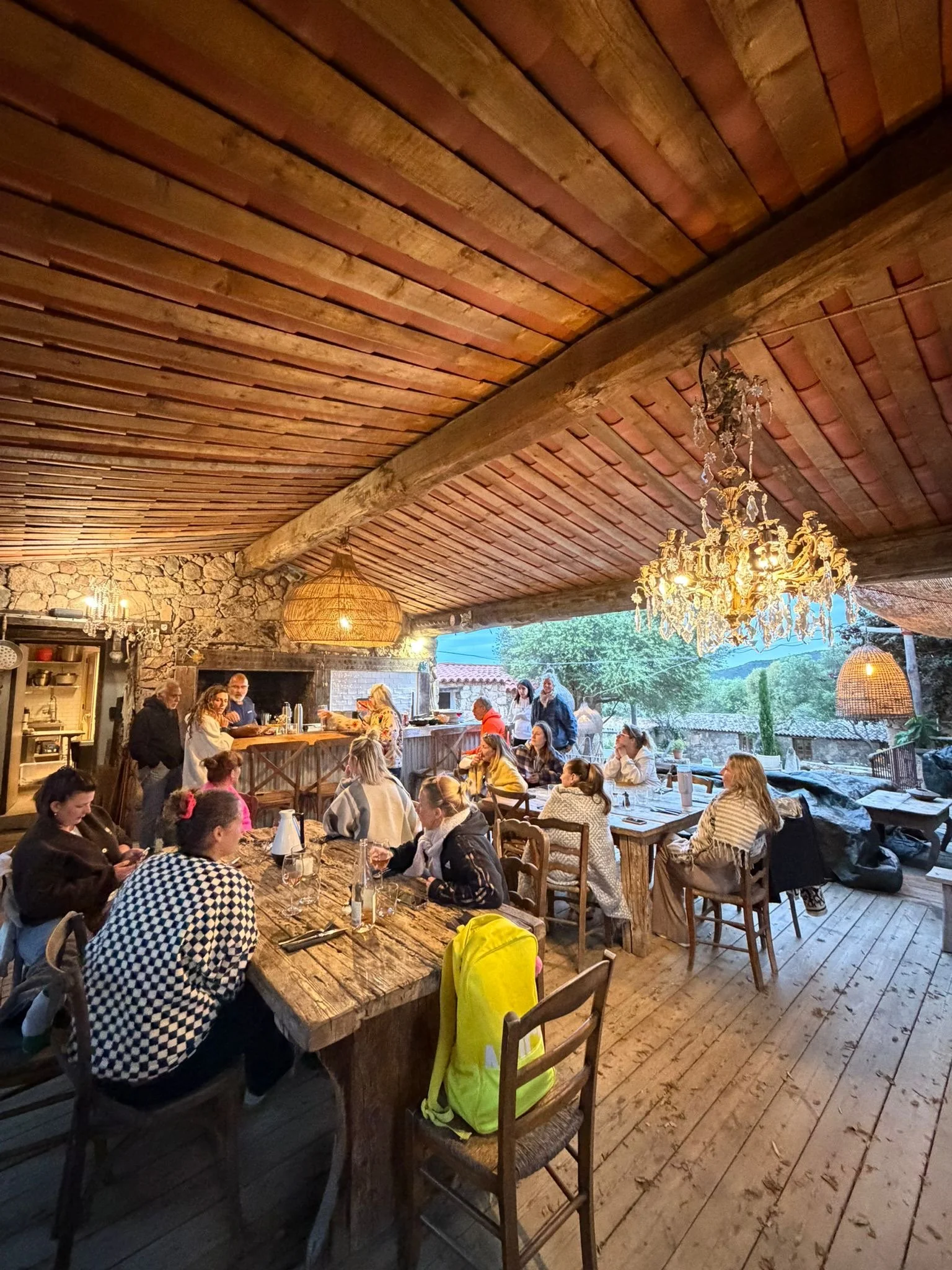 Une scène de repas dans une ferme ou un restaurant, avec des personnes assises autour de longues tables en bois, à l'intérieur d'une structure en bois avec des luminaires en cristal et des lampes en osier, et une vue sur la nature extérieure.