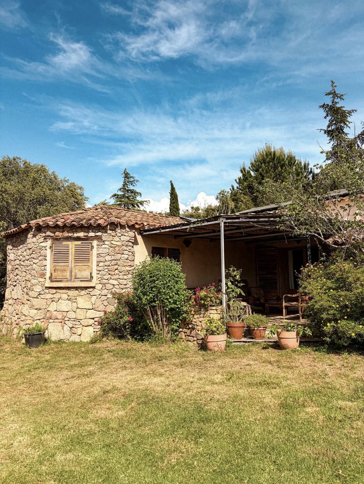 Maison de campagne en pierre avec un toit en tuiles rouge et une terrasse pleine de plantes en pot, entourée de jardin et de grands arbres, sous un ciel bleu avec quelques nuages