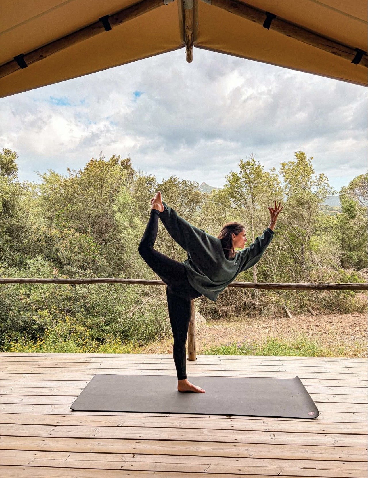 Une femme pratique le yoga en position d'équilibre sur un tapis noir, sous une structure en bois, avec un arrière-plan de forêt et un ciel nuageux.