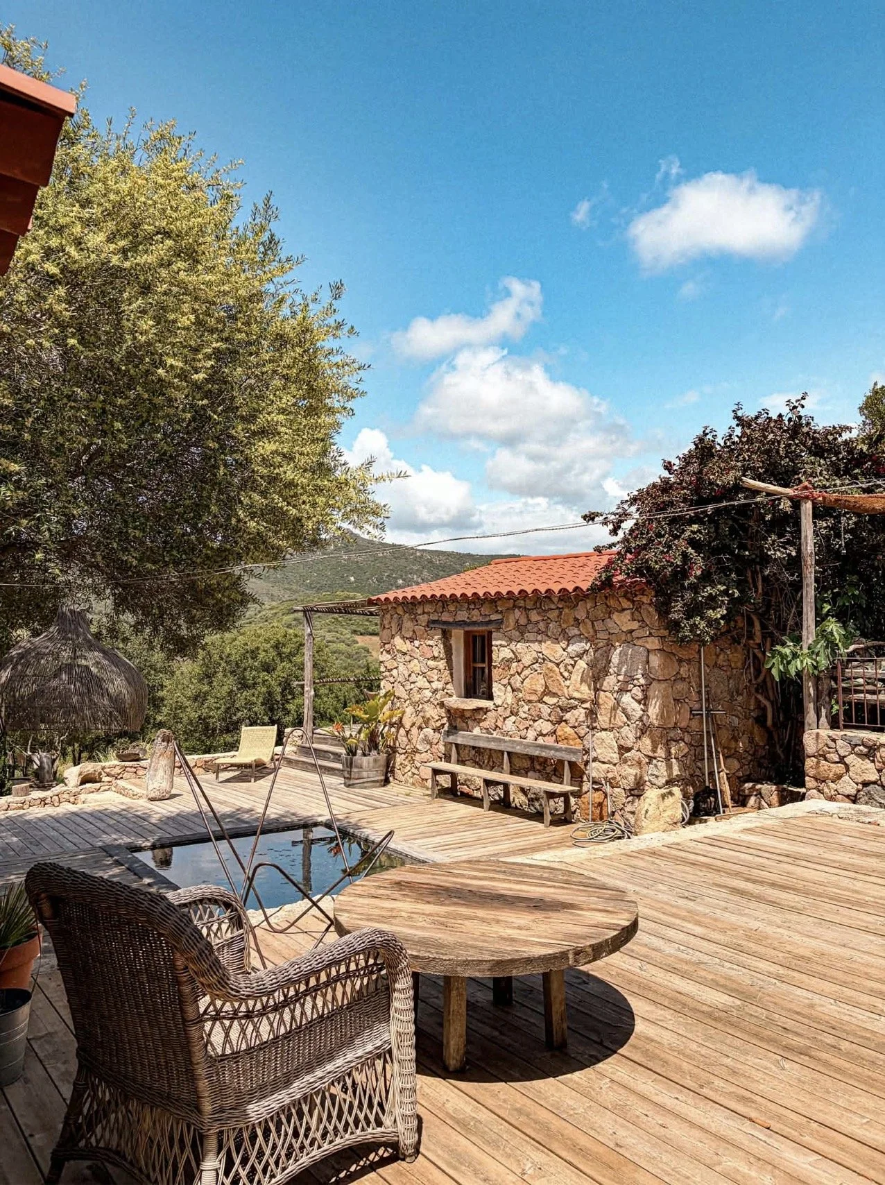Maison en pierre avec toit en tuiles rouges, terrasse en bois, piscine, mobilier extérieur dont chaise en rotin et table en bois, arbres et ciel bleu avec nuages.