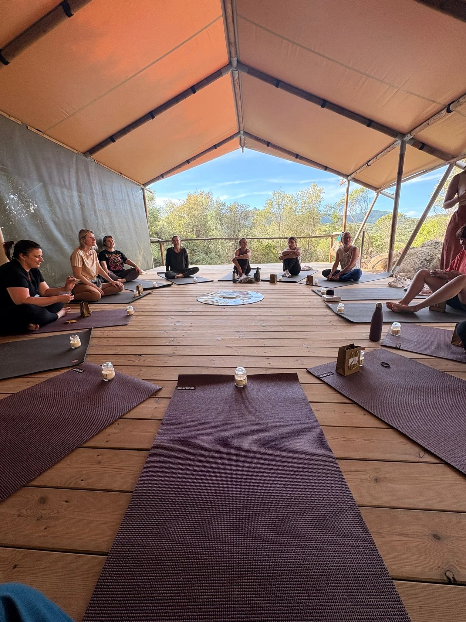 Groupe de personnes assises en cercle sur des tapis de yoga dans une cabane en bois avec vue sur la nature et un ciel bleu.