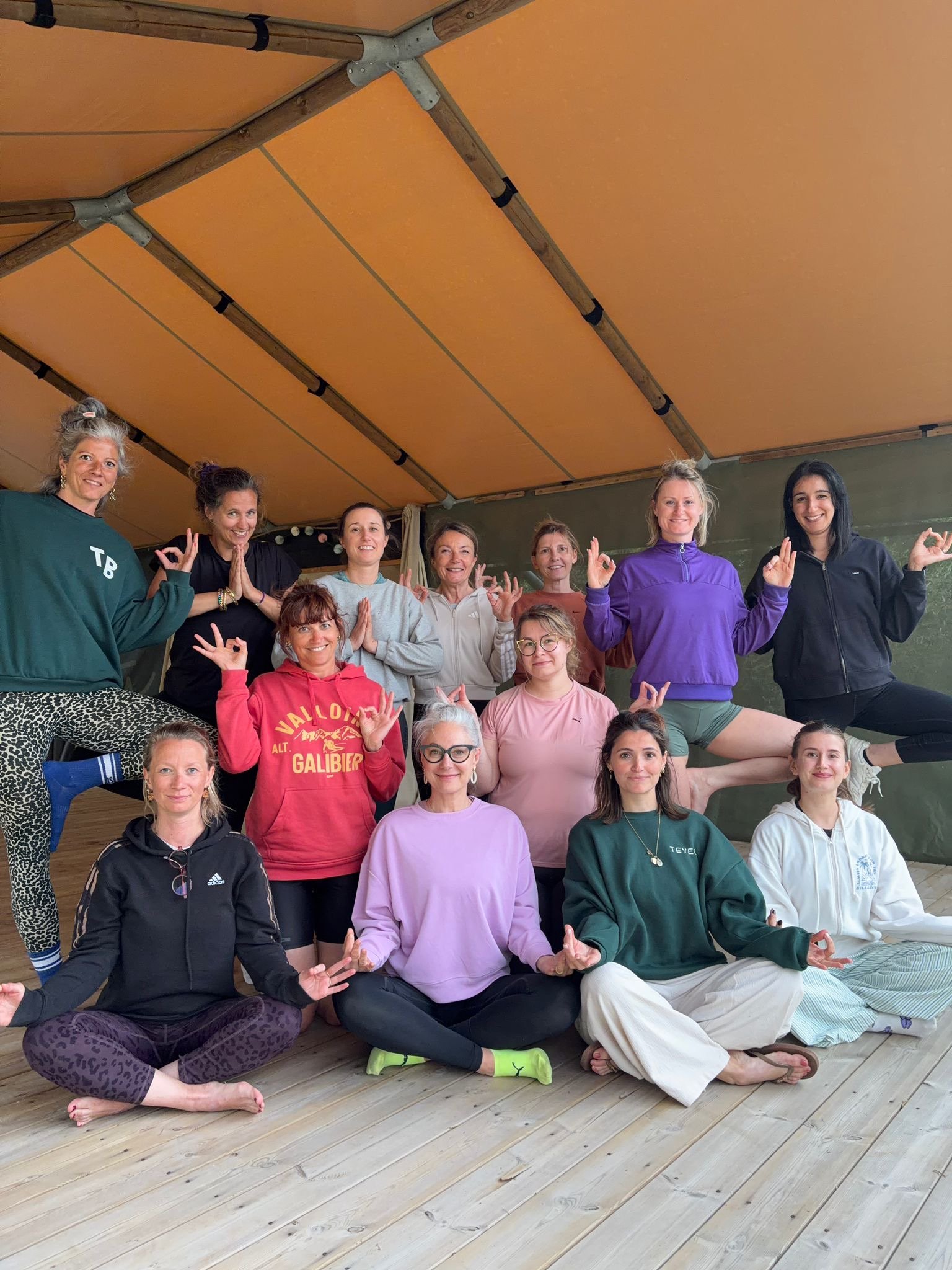 Groupe de femmes pratiquant la méditation ou le yoga en intérieur, certaines en position de lotus, faisant des gestes de méditation avec les doigts, dans une ambiance conviviale.