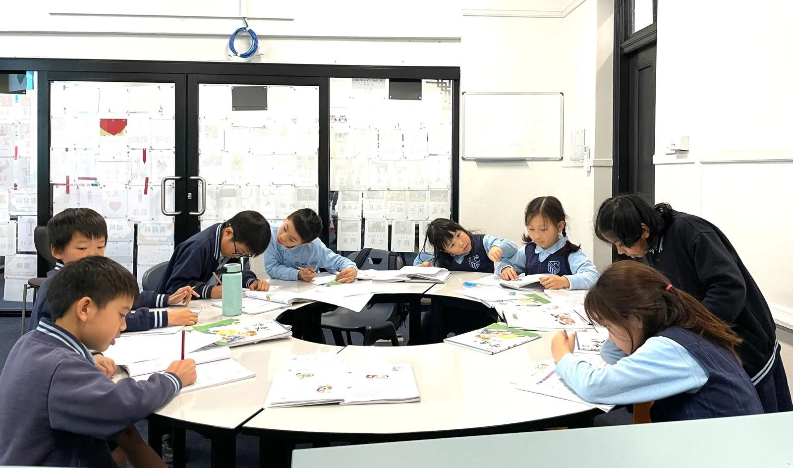 A classroom with eight children and one adult, all engaged in reading or writing at a large circular table. The children appear to be students, and the adult looks like a teacher. The room has floor-to-ceiling windows with paper notes or artwork tape