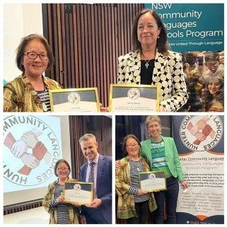 Three women and one man at a community languages program award event, holding certificates, with banners and a logo in the background.