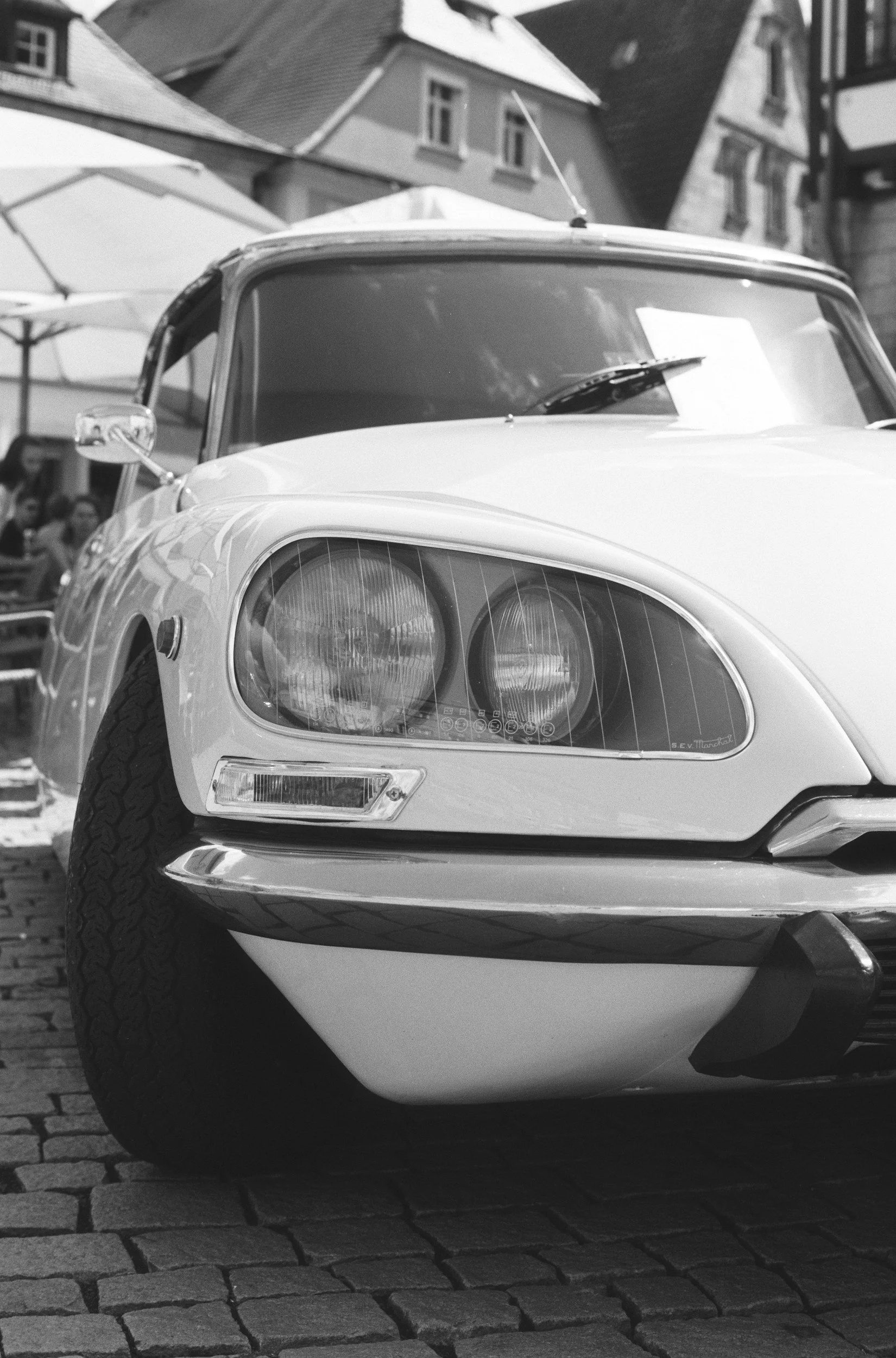 A vintage sports car parked on a cobblestone street, with buildings and umbrellas in the background, in black and white.
