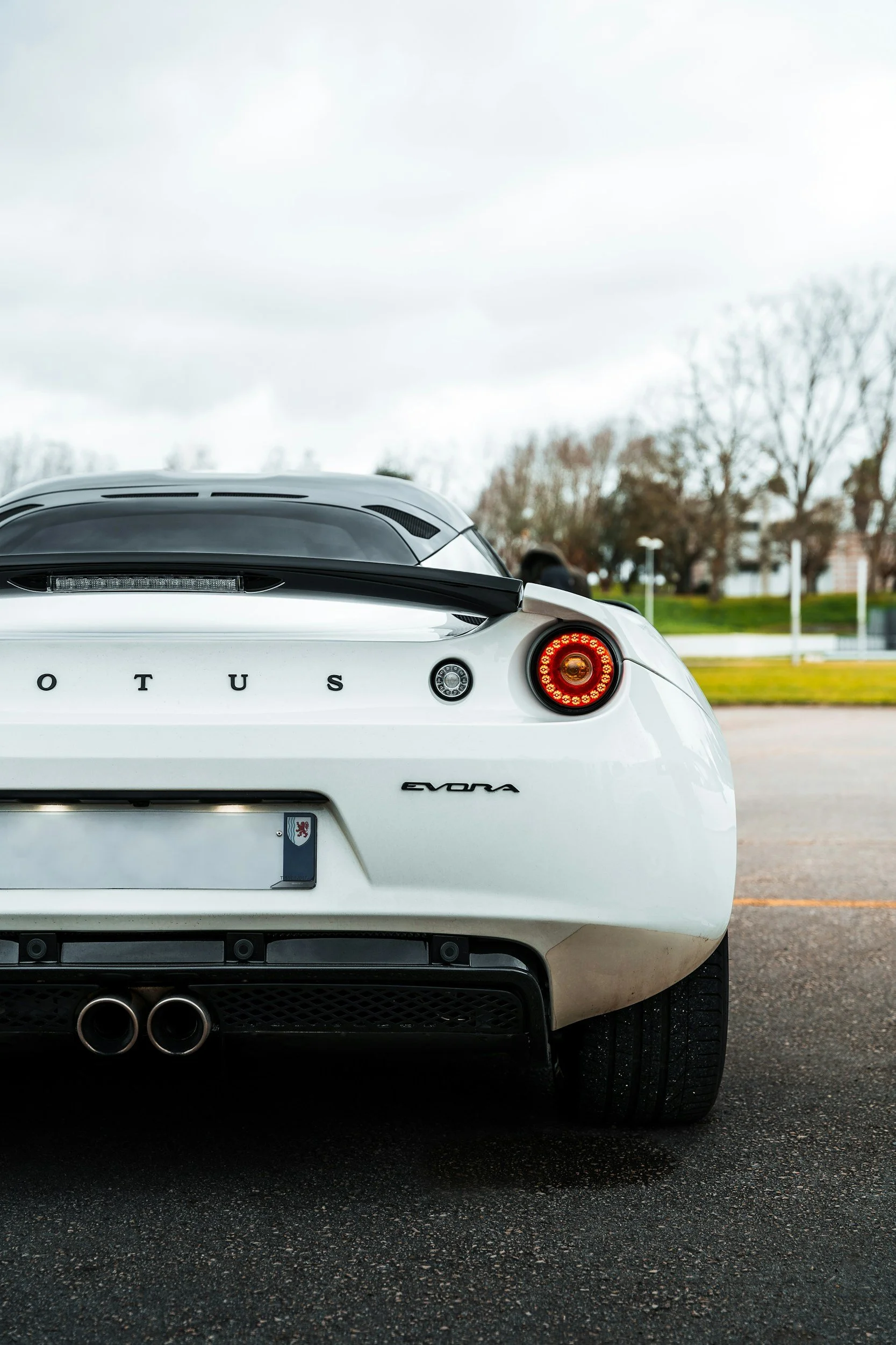 Rear view of a white Lotus Evora sports car parked on a paved lot with trees and grass in the background.