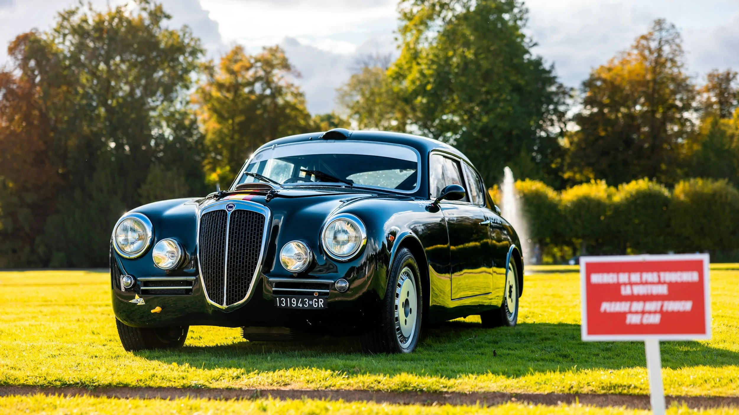 A vintage black car parked on a grassy area with a red warning sign in front that says 'Please do not touch the car.' The background features trees and a fountain under a cloudy sky.