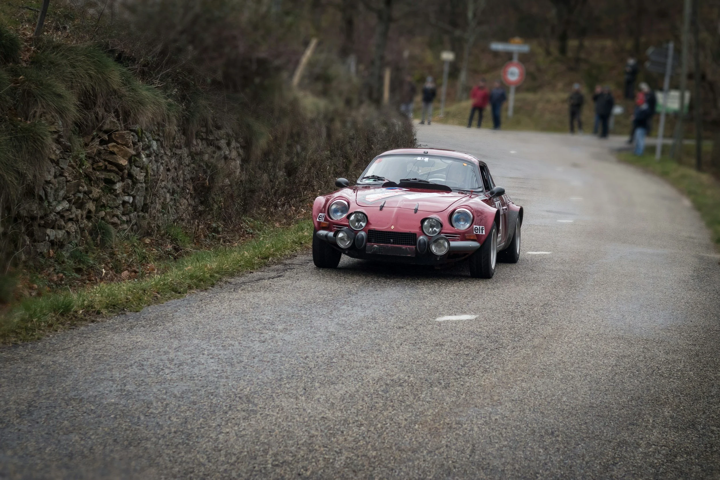 A classic red rally car with multiple headlights on a winding rural road, with spectators in the background.