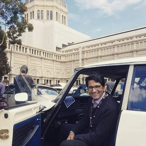A smiling man wearing glasses and a dark sweater sitting in an open car on a city street with historic architecture behind him.