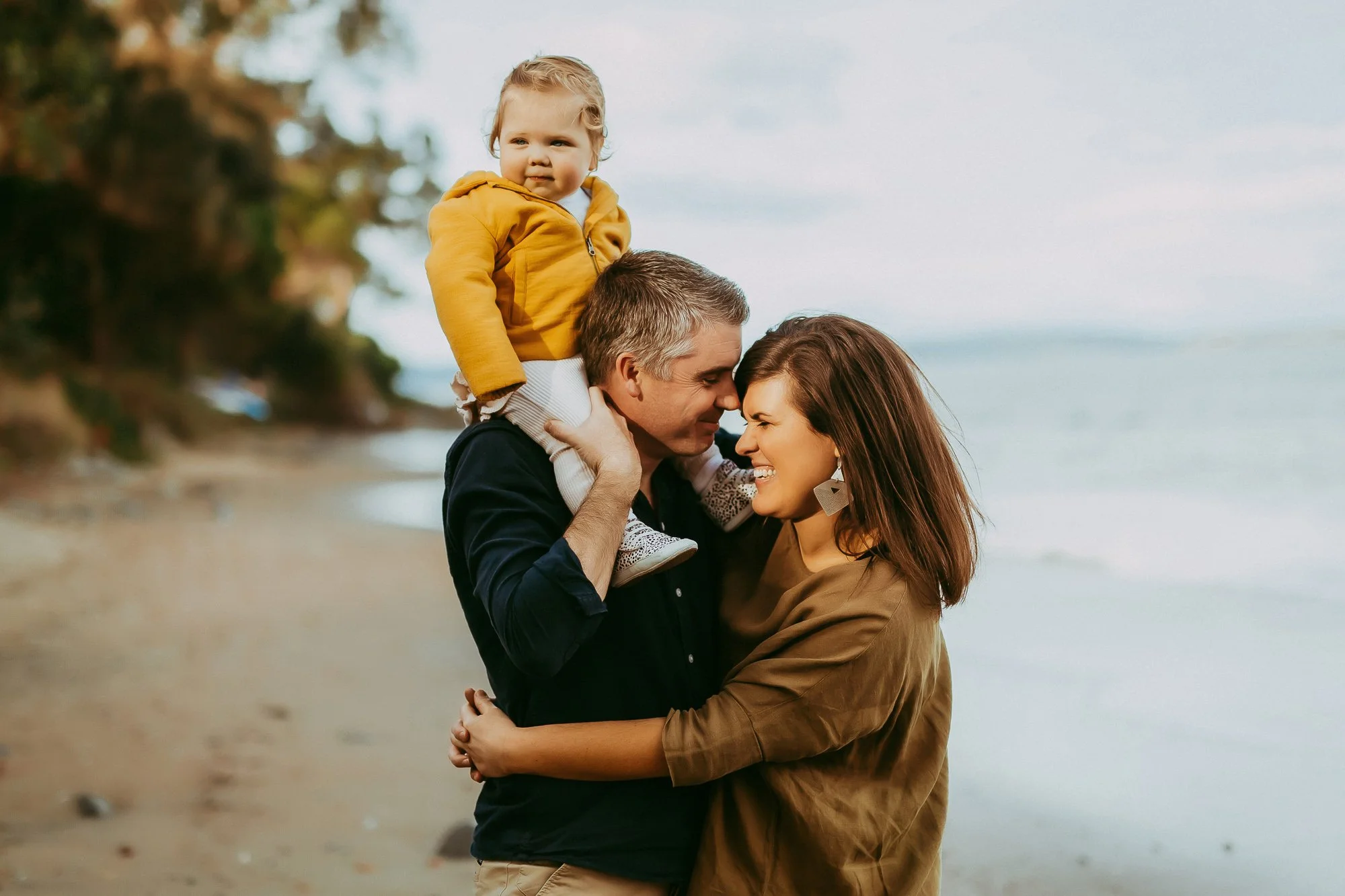 A family of three enjoying a moment together on a beach, with a man holding a young girl on his shoulders and a woman embracing him, all smiling at each other. Greater Hobart family photosession with relaxed approach.