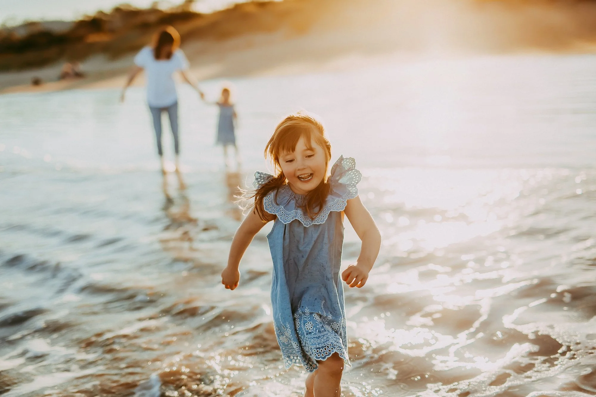 A young girl running and playing in the shallow water at the beach, with a woman and another child in the background during sunset.