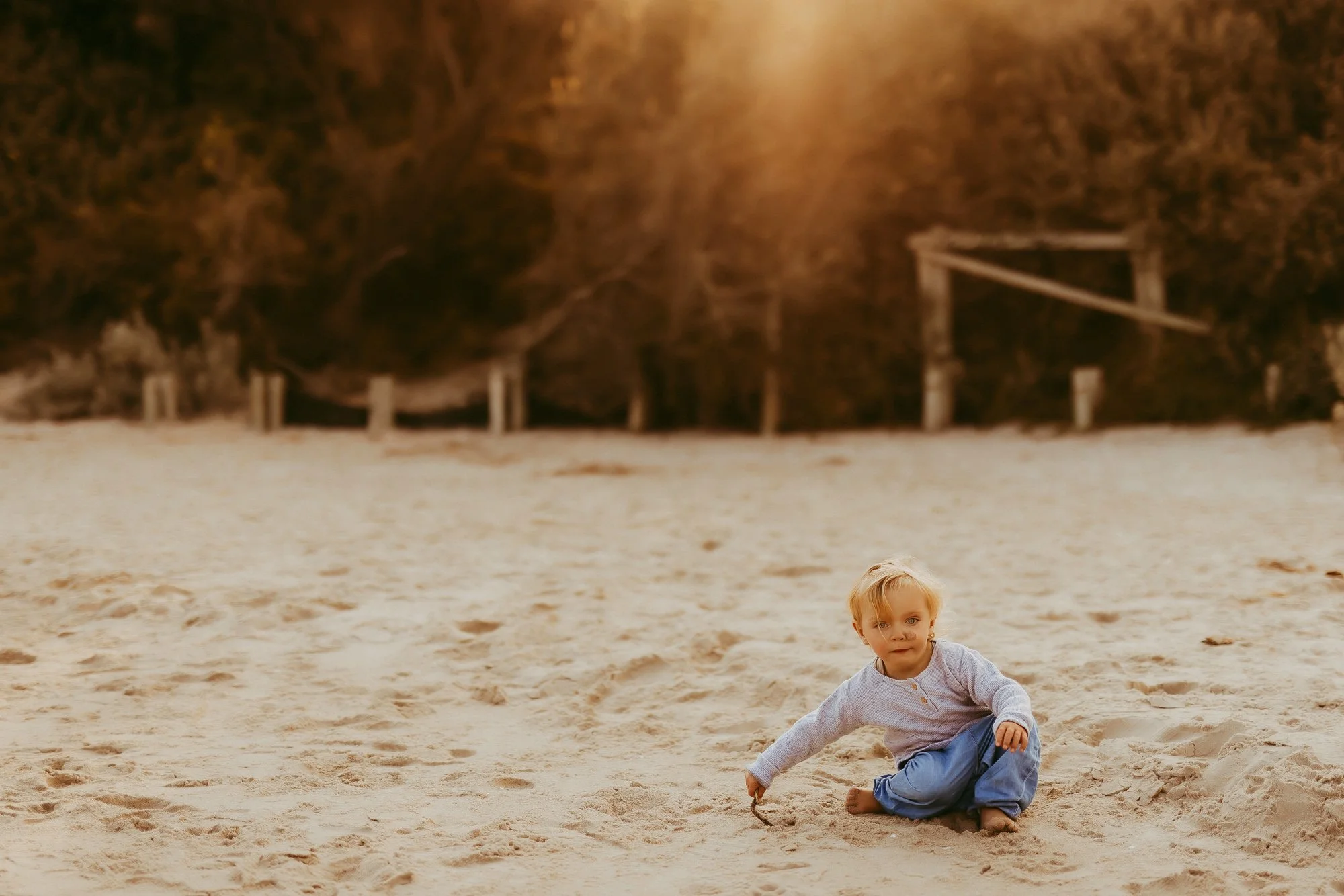 A young boy with blonde hair sitting on a sandy beach, digging in the sand. In the background, there are trees and a wooden structure.